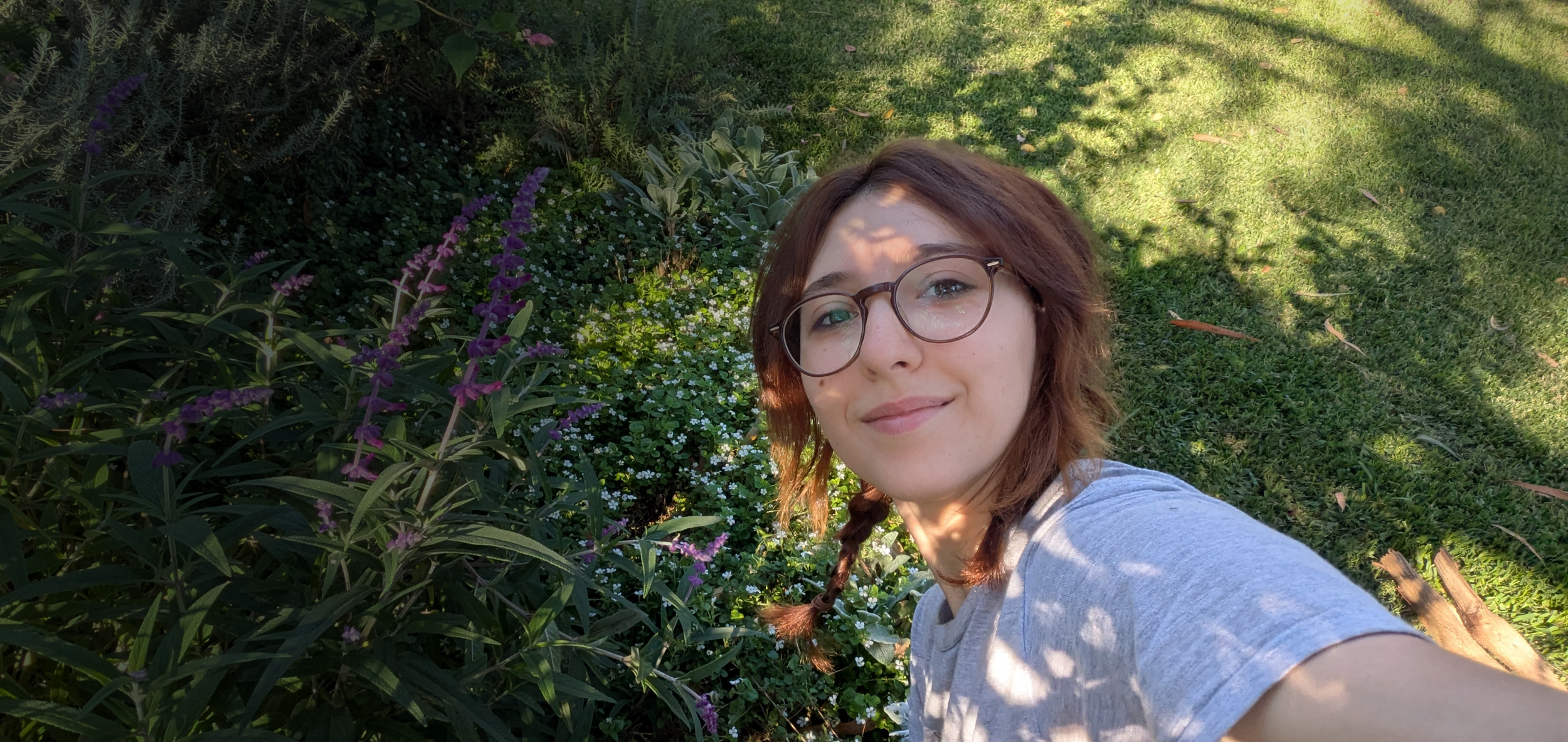 A young woman with glasses and braided hair taking a selfie outdoors in a garden with purple and white flowers and green grass.