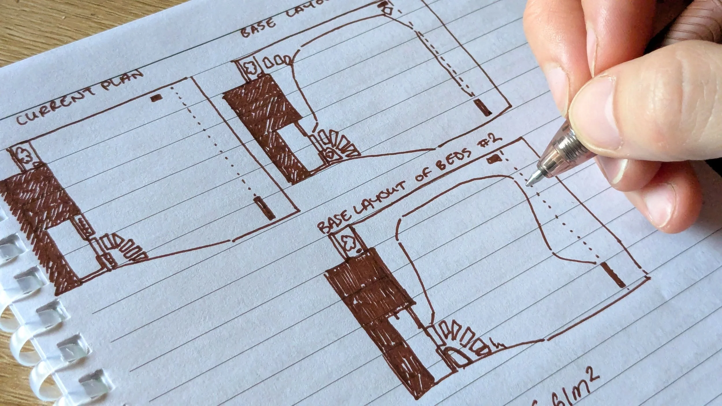A hand drawing a floor plan with a brown pen on lined notebook paper, showing rooms and labels such as "Current Plan", "Base Layer of Beds", and "Base Case Cargo".