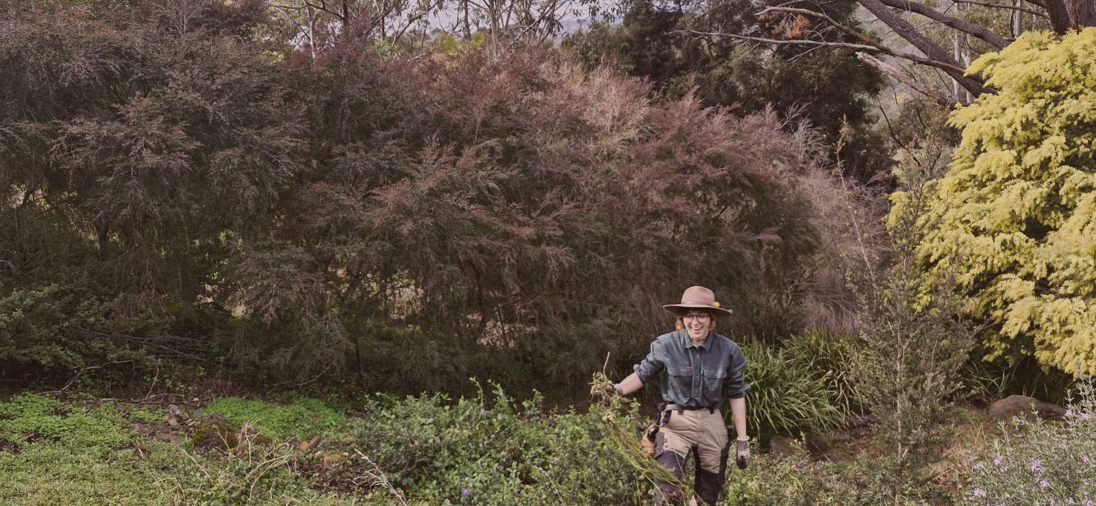 A woman in outdoor gardening attire with a wide-brimmed hat, glasses, gloves, and khaki pants stands in a lush garden surrounded by trees and plants, holding a small plant or branch.