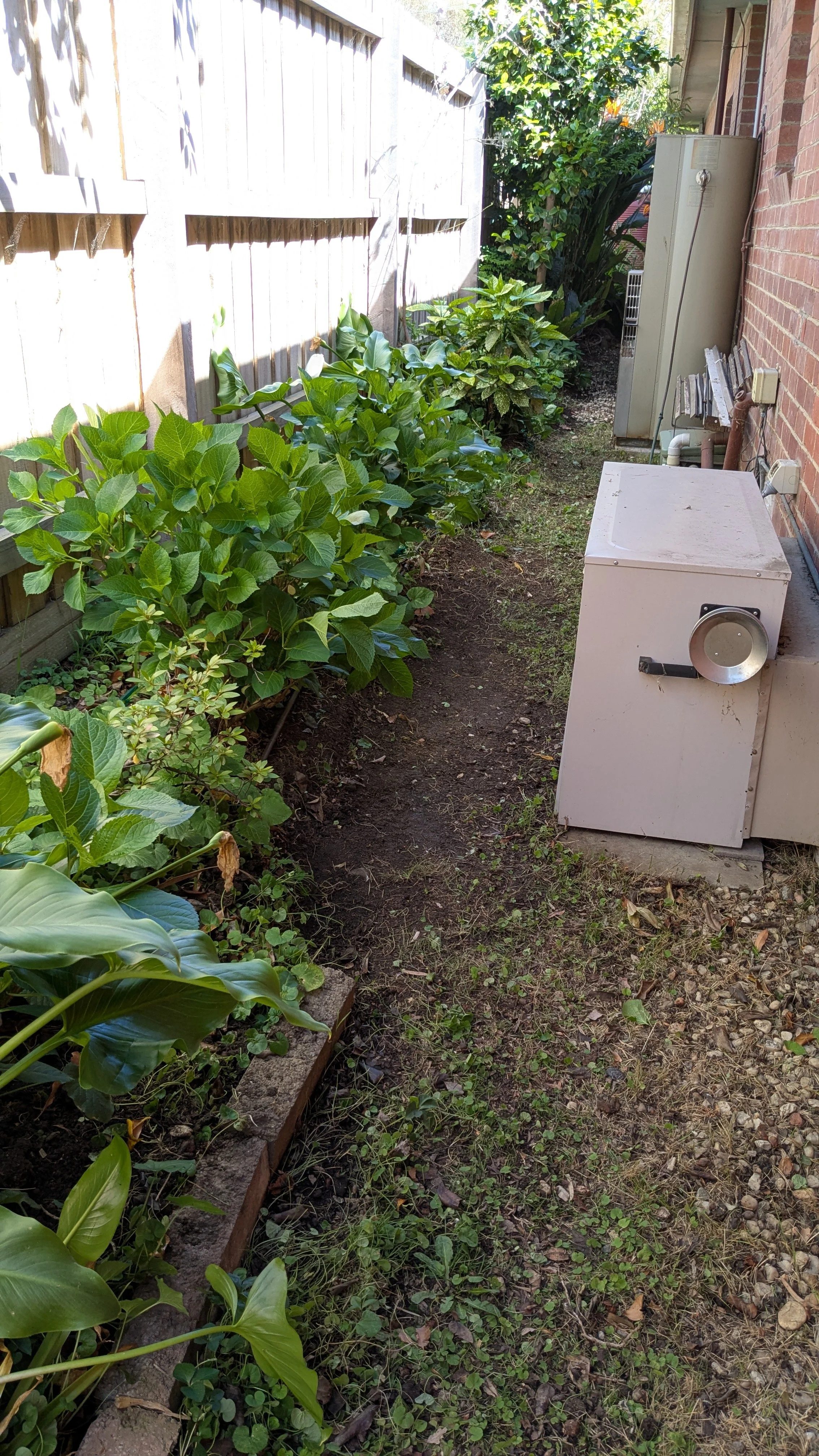 A narrow backyard garden with green plants along a wooden fence on the left and a brick wall on the right. There is a white utility box in the foreground, and an air conditioning unit and electrical components along the brick wall. The ground is a mi