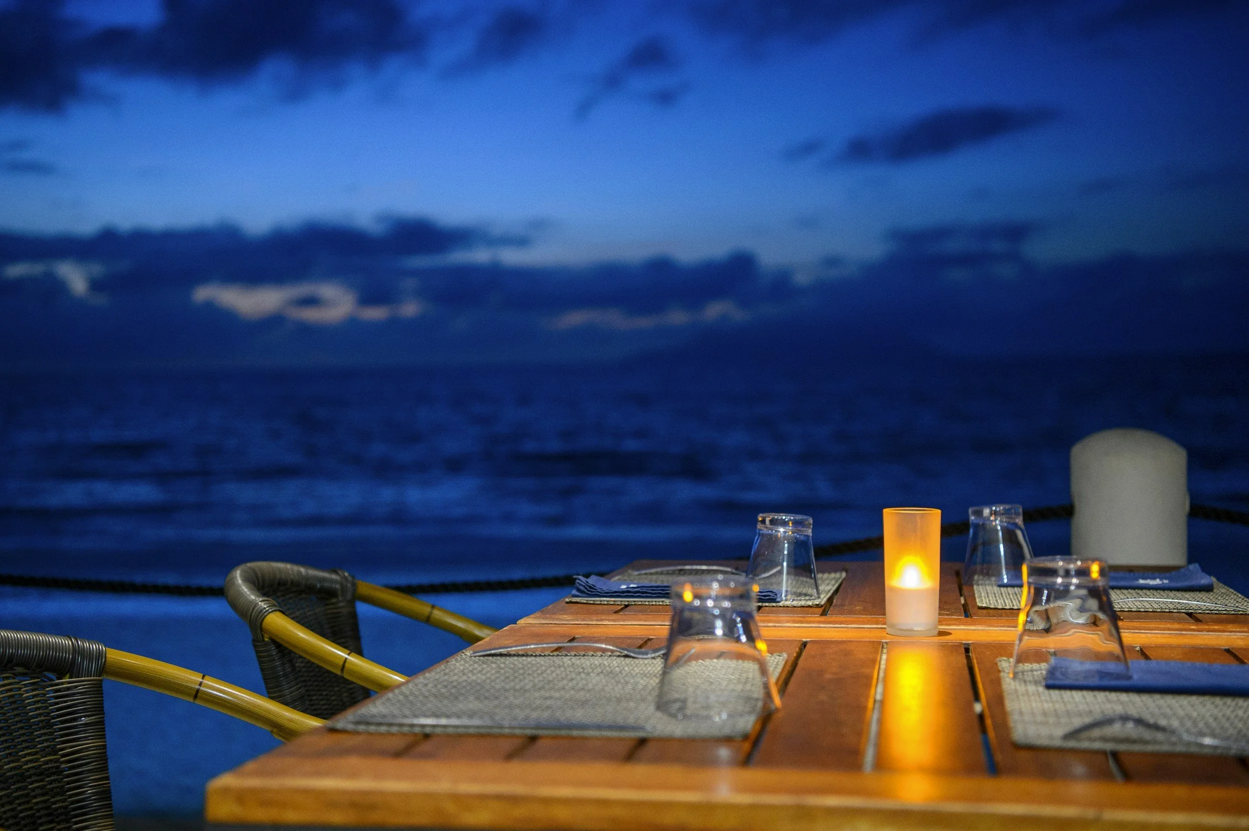 An outdoor dining table set with four placemats, glasses, and a candle, overlooking the ocean at twilight with dark clouds in the sky.
