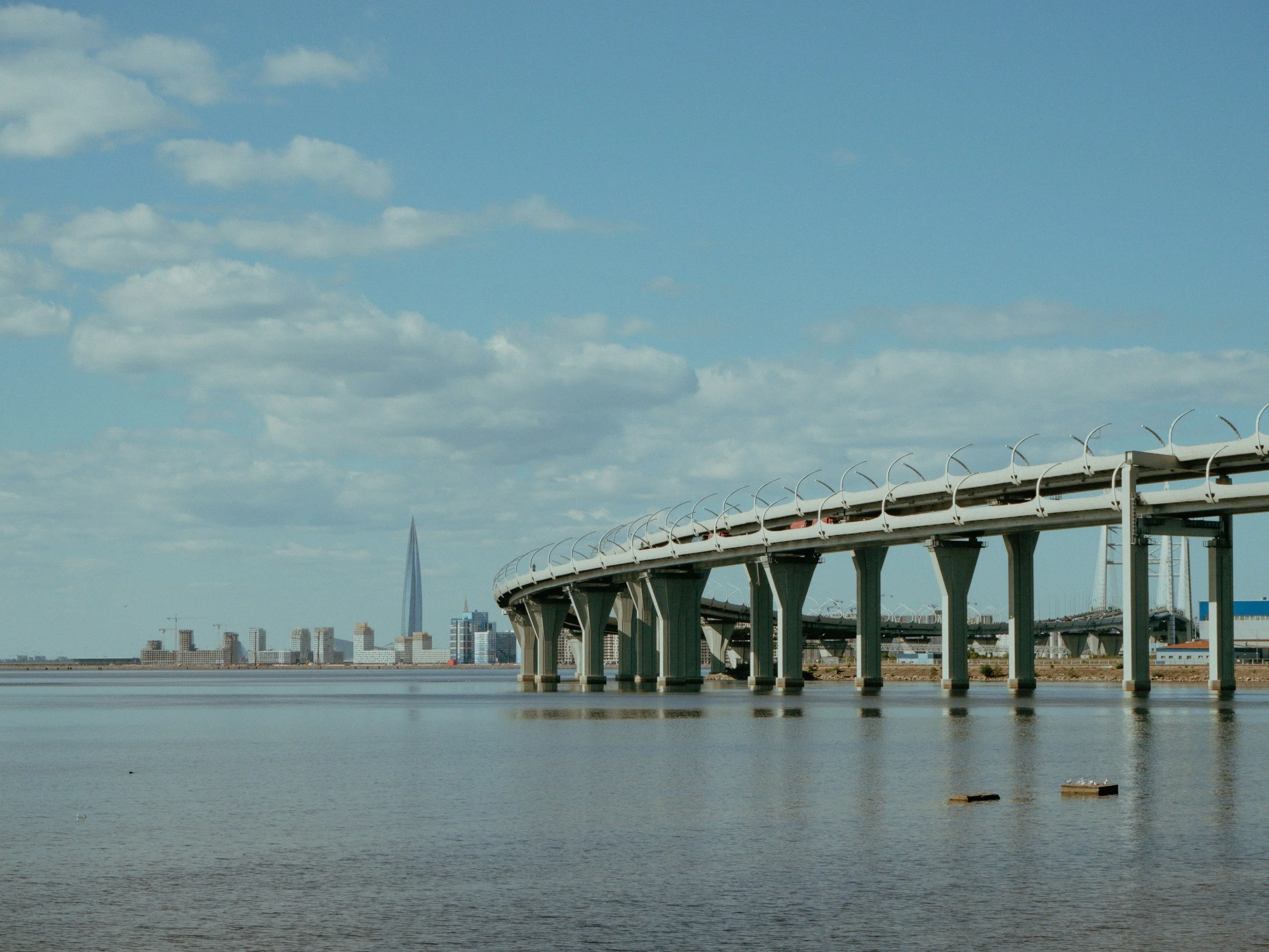 Photo of a bridge over water with a city skyline in the background, including a tall, slender building and a ferris wheel, under a partly cloudy sky.
