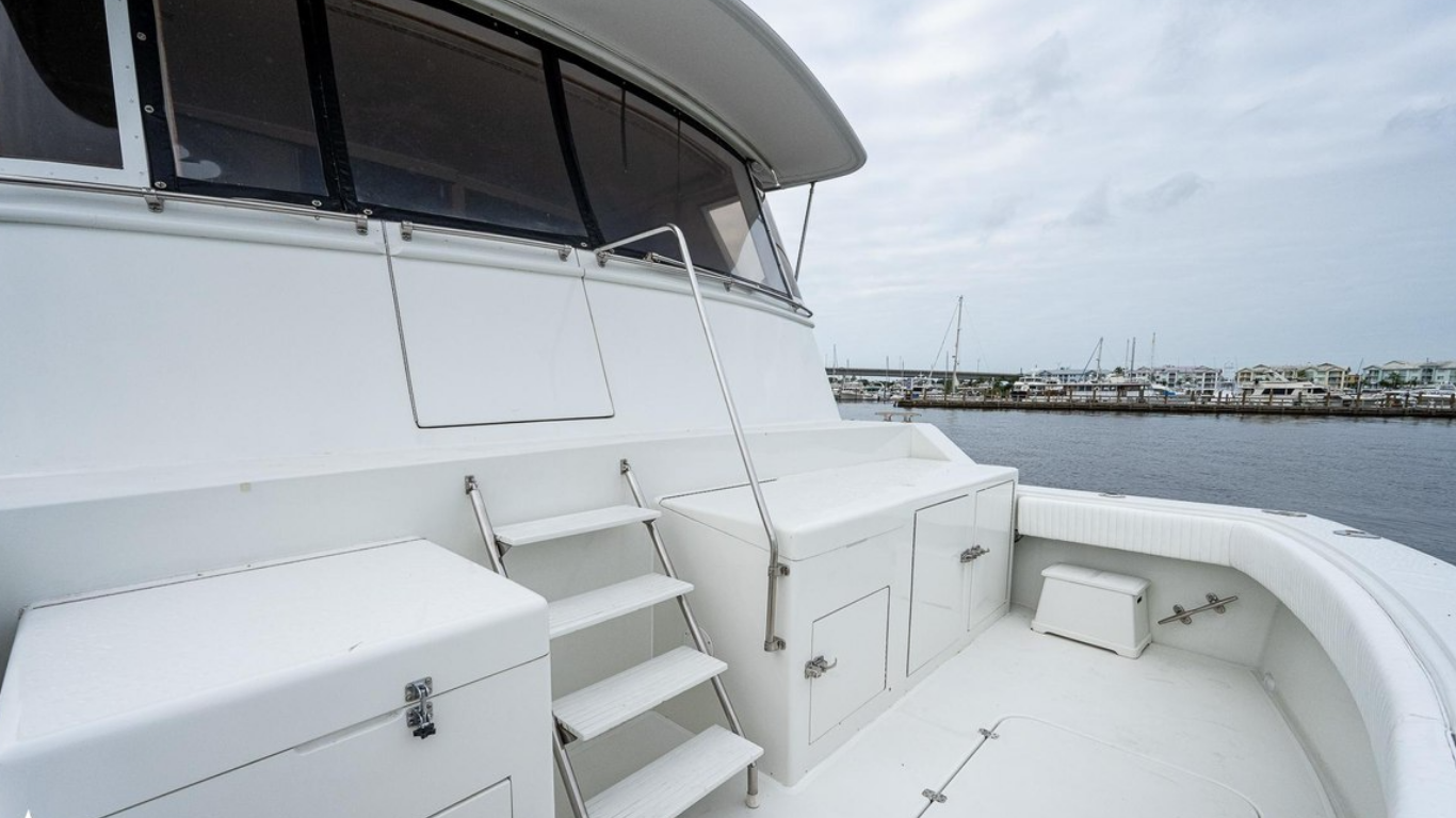 View of the deck of a luxury white yacht docked at a marina, with several boats and houses in the background under a cloudy sky.