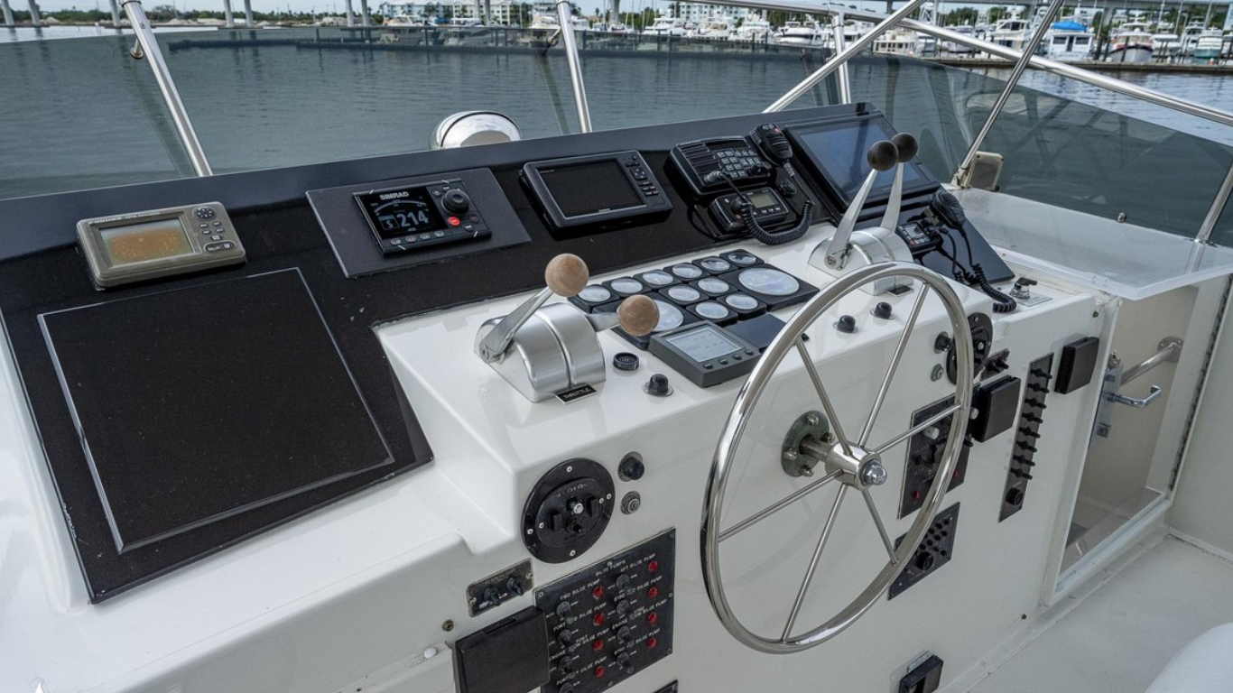 Control panel of a boat with navigation equipment, gauges, communication devices, and a metal steering wheel, with moored boats and water in the background.