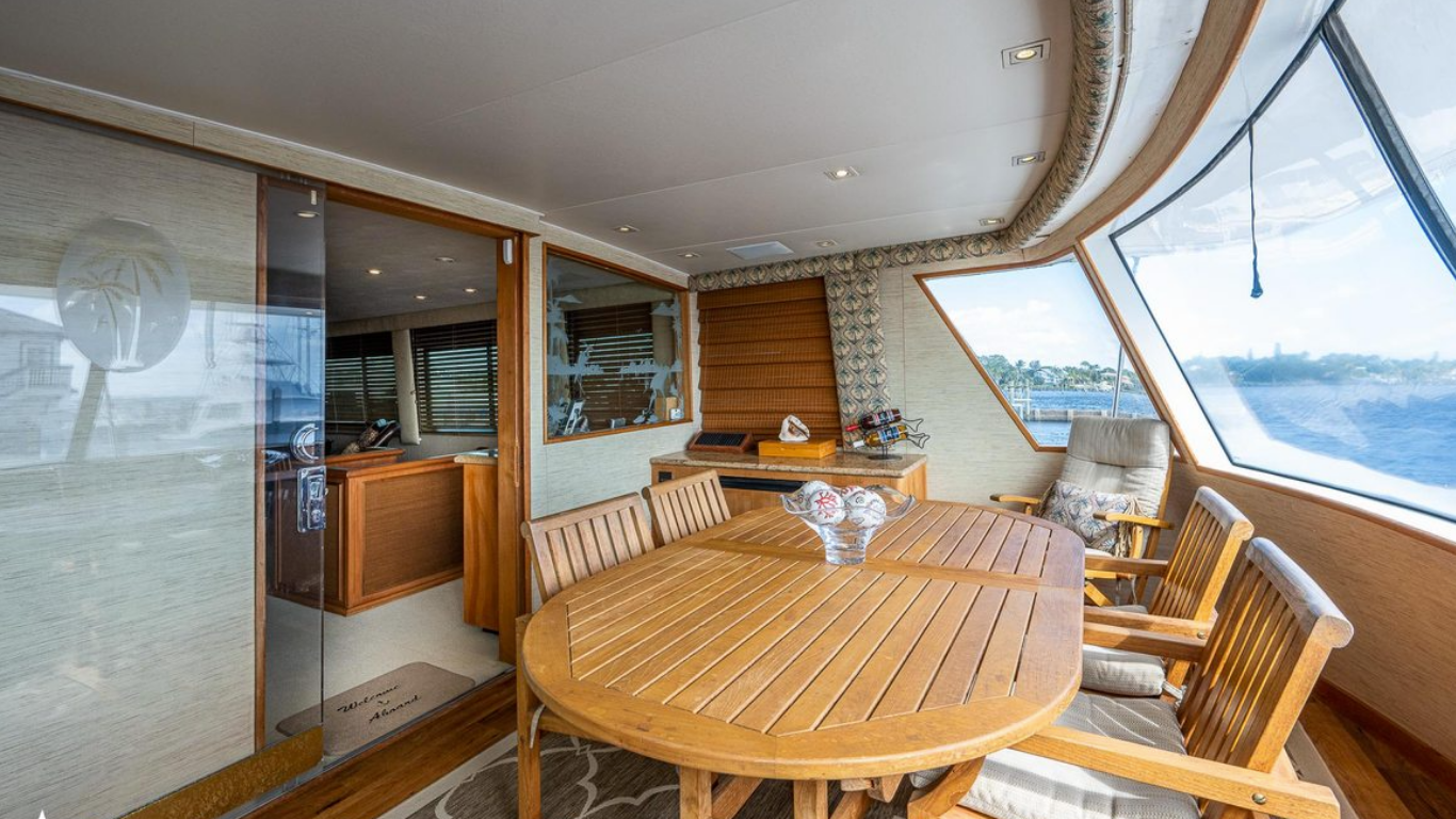 Dining area with wooden table and chairs inside a boat or yacht, large windows showing water and sky outside, decorative bowl on table, window blinds, and seating area with cushions