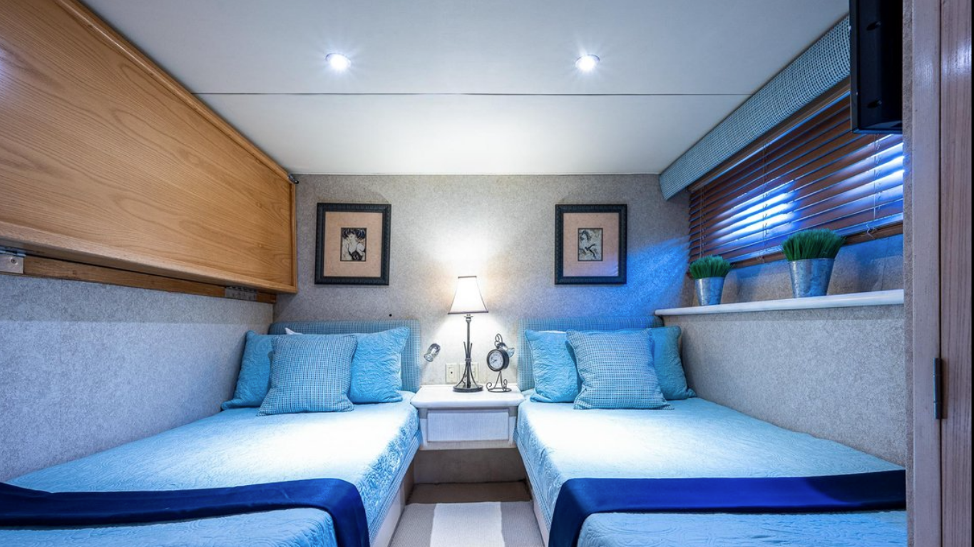 A cozy bedroom with two twin beds covered in light blue bedspreads and matching pillows, a small nightstand with a lamp and decorative items, two framed pictures on the wall, a window with wooden blinds and potted plants on the windowsill.