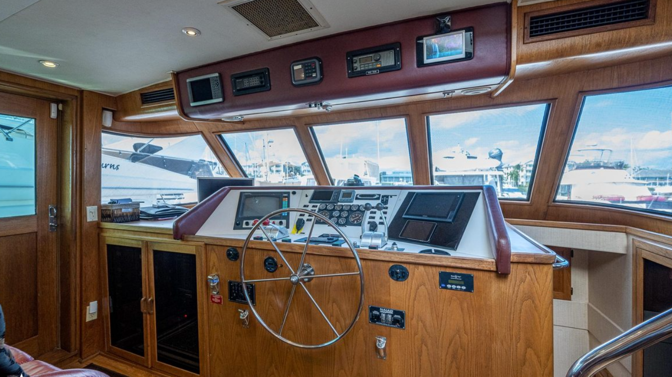 The image shows the interior of a boat's helm station with a large wooden steering wheel, navigation and control panels, and multiple large windows providing a view of the marina with several boats docked outside.