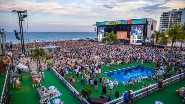 Pink and white ornate building with arched balconies and decorative architectural details, with a palm tree branch visible in the top right corner and a partly cloudy sky in the background.