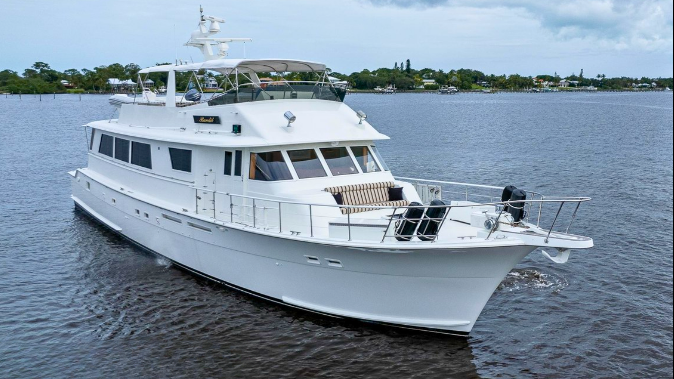 A white yacht cruising on a body of water with buildings and trees in the background under a partly cloudy sky.