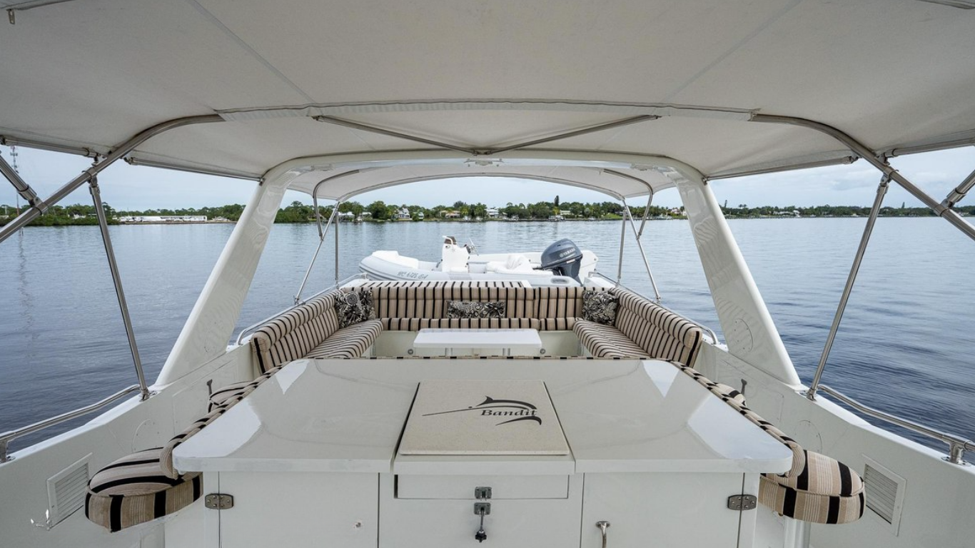 View from the deck of a boat showing seating area with striped cushions, a small table, and a boat with an outboard motor on calm water with houses and trees in the background.