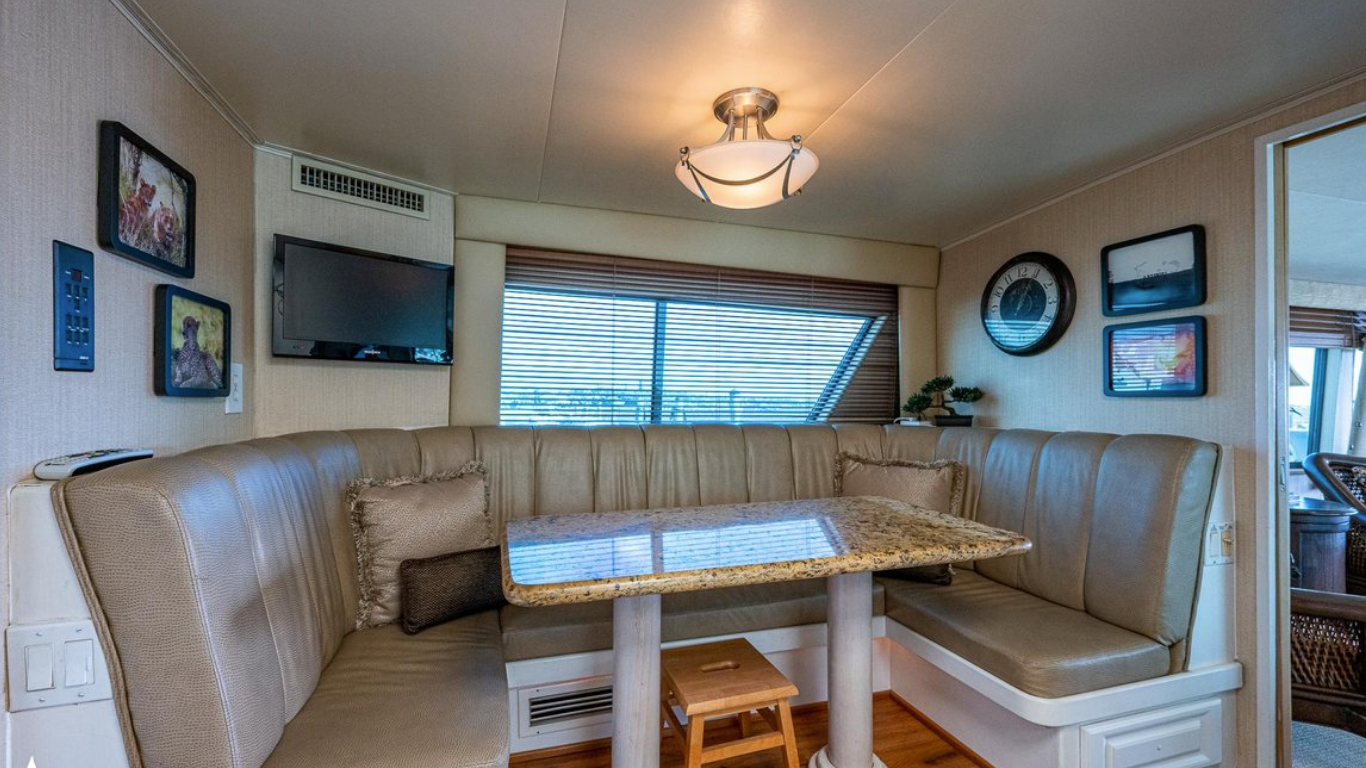 Interior view of a living space with a corner beige leather booth, a granite dining table, wall-mounted TV, framed pictures, a clock, and a window with blinds.