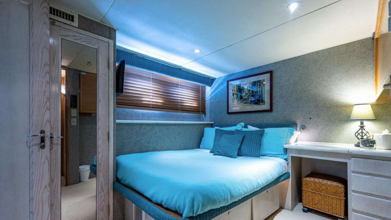 A cozy hotel room with a neatly made bed covered in turquoise bedding, a small nightstand with a lamp, a wall-mounted TV, a framed picture on the wall, and a view into a bathroom with a toilet visible.