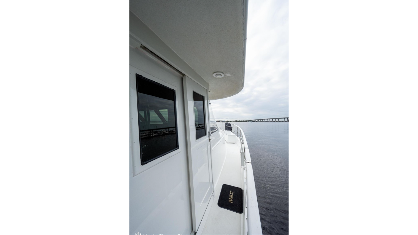 View from a boat showing a white exterior wall with two windows and a door, a black mat with the word 'BANDIT,' water, and a bridge in the distance under a partly cloudy sky.