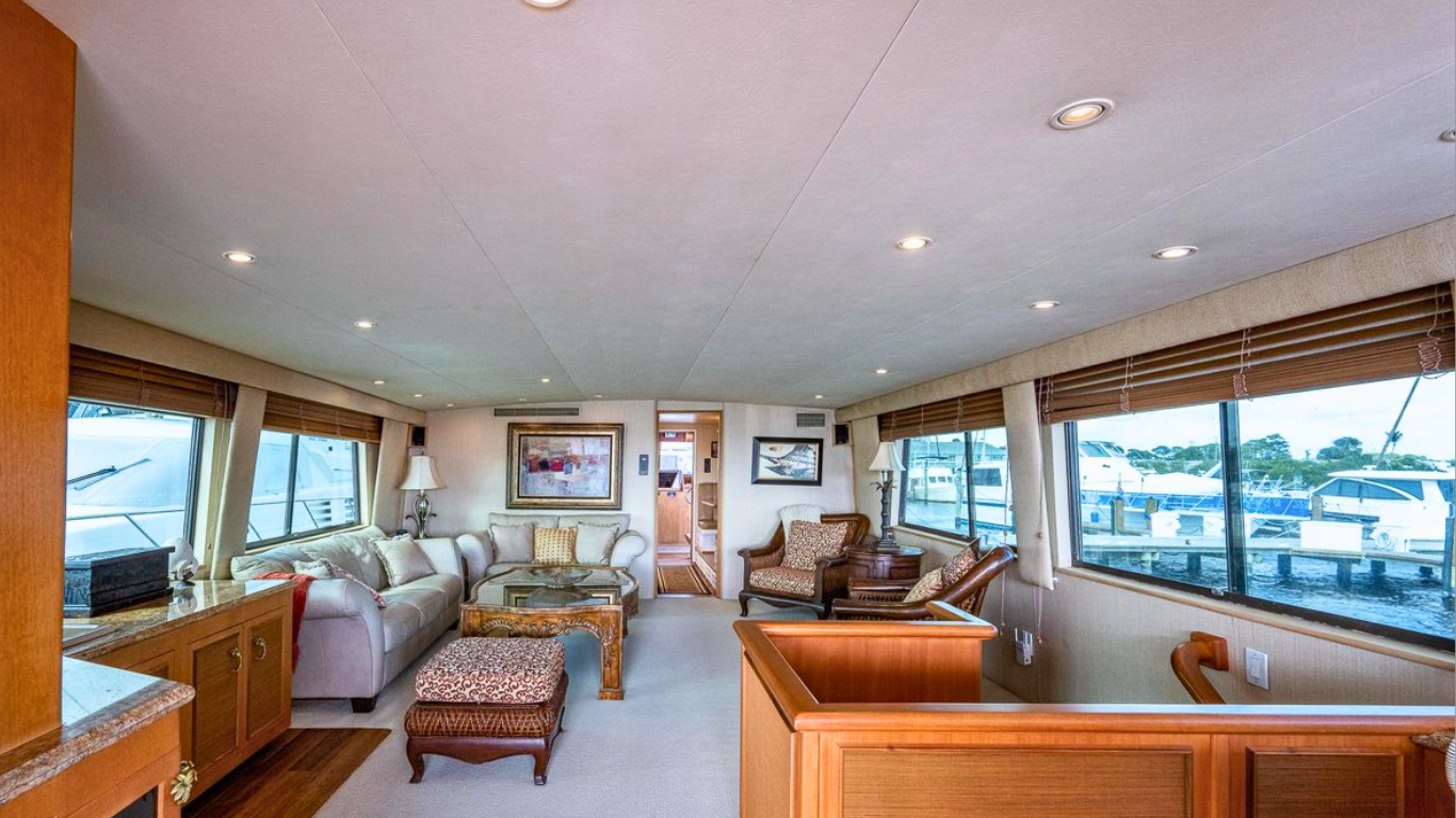 Living room with large windows showing boats docked outside, beige sofas, armchairs, artwork on the walls, and wooden furniture.