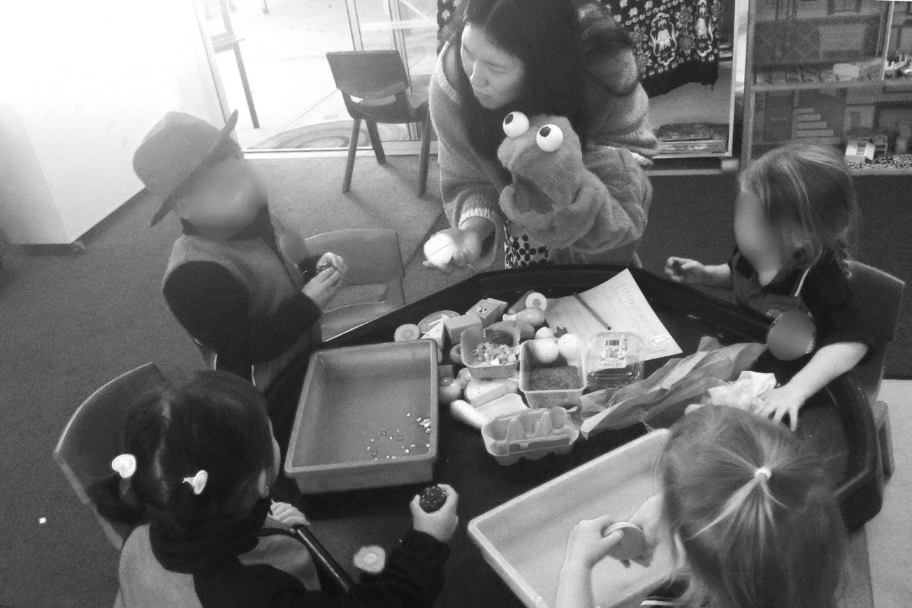 A woman holding a puppet in a classroom with four children around a table filled with toys and art supplies.