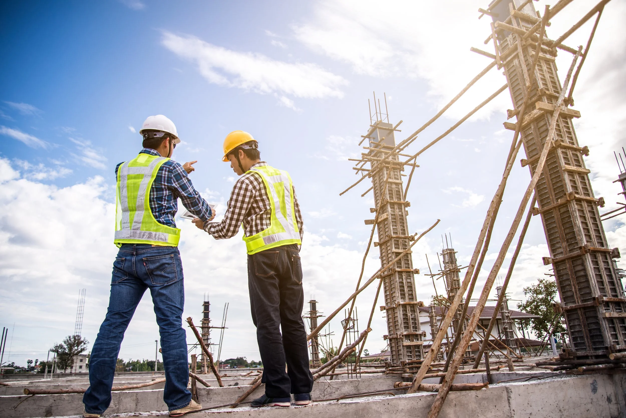Two construction workers wearing safety vests and helmets, standing on a building site with vertical concrete columns and wooden supports, under a partly cloudy sky.