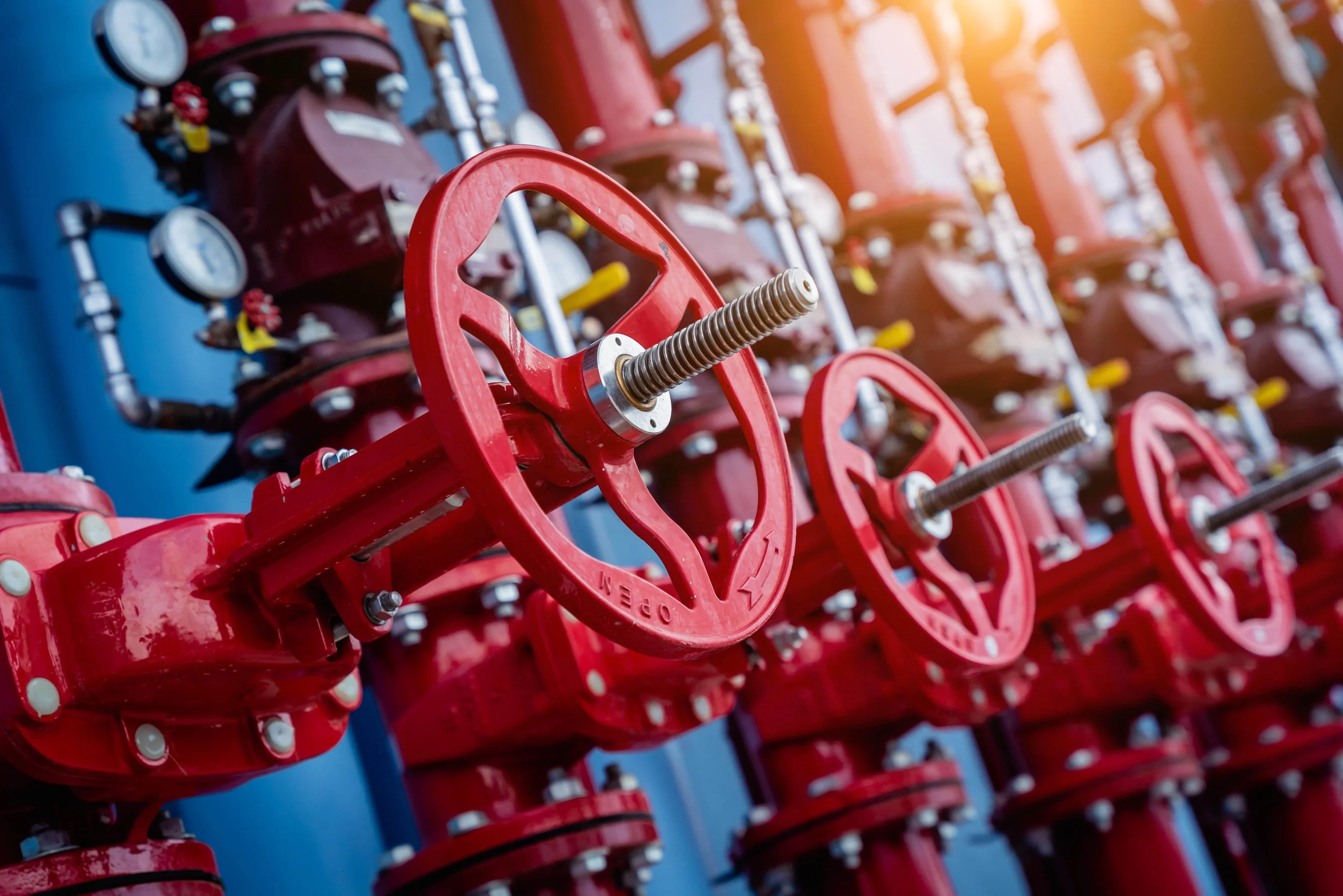 Red industrial valve wheels and gauges on a blue wall, with sunlight in the background.