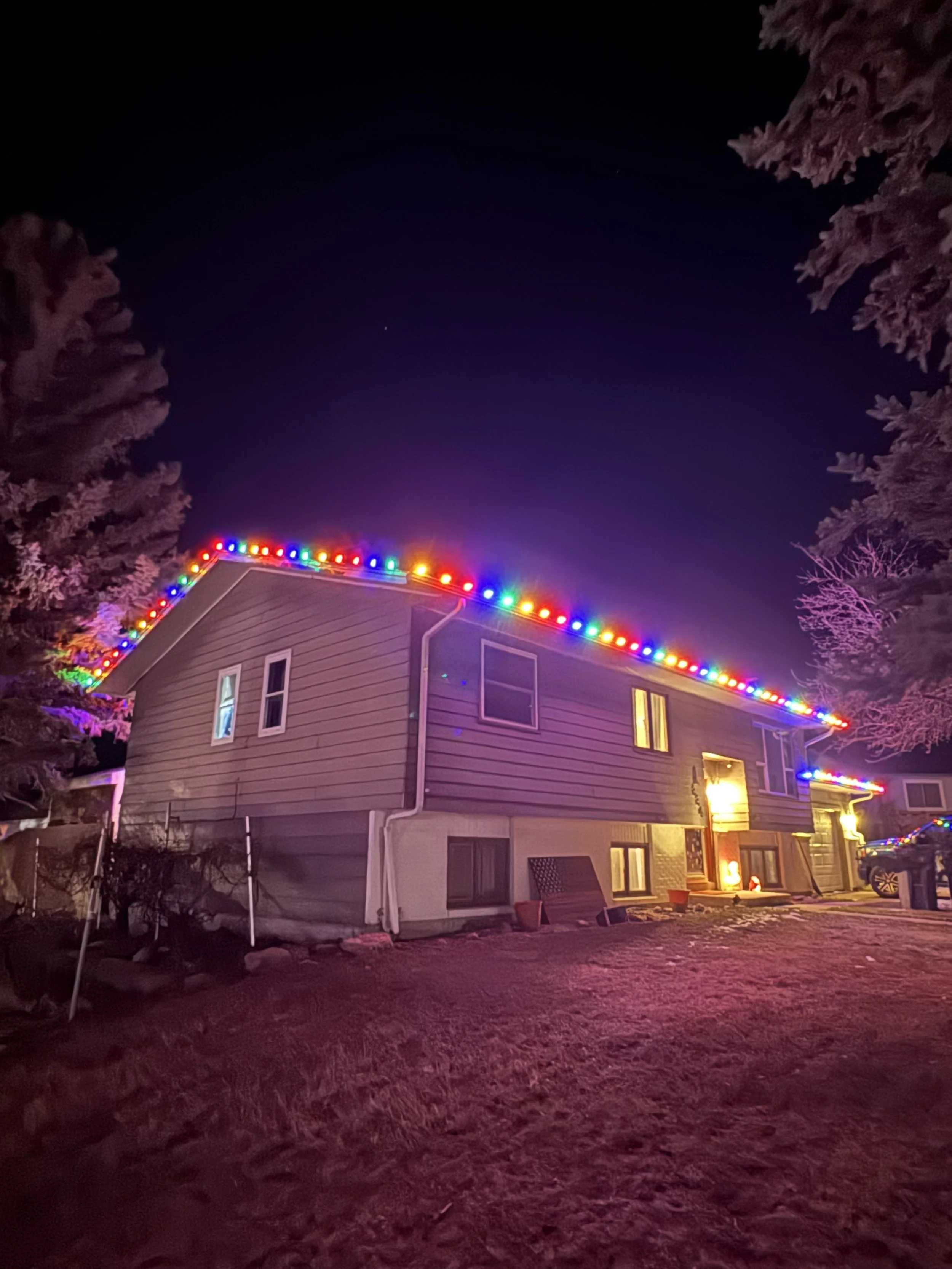 A house at night with colorful Christmas lights along the roof edge, surrounded by leafless trees. The scene is illuminated by indoor lights and outdoor lighting, with a clear dark sky overhead.