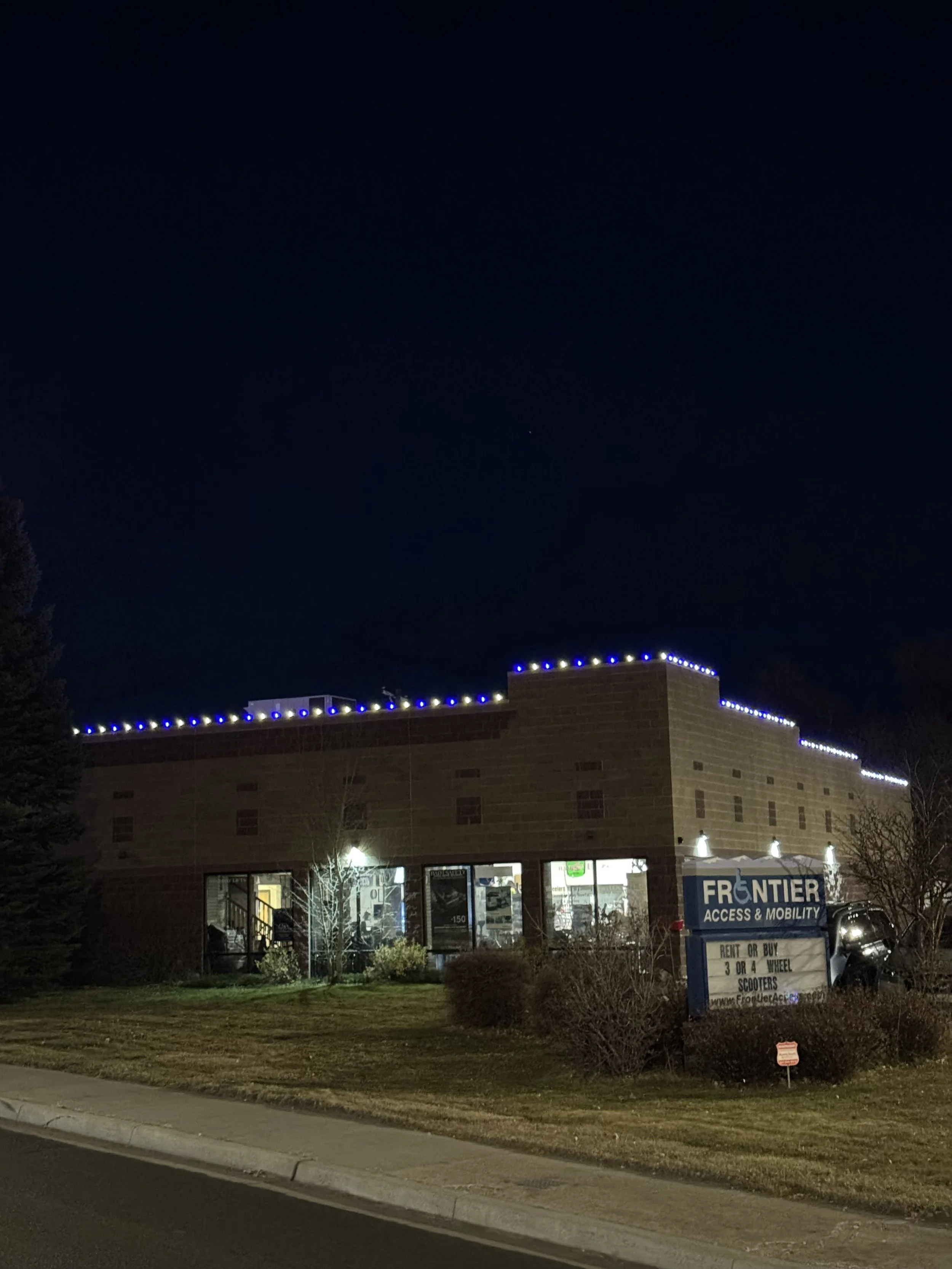 Night view of a building with blue and yellow decorative lights on the roof, store windows on the ground floor, and a sign for Frontier Access & Mobility in front of the building.