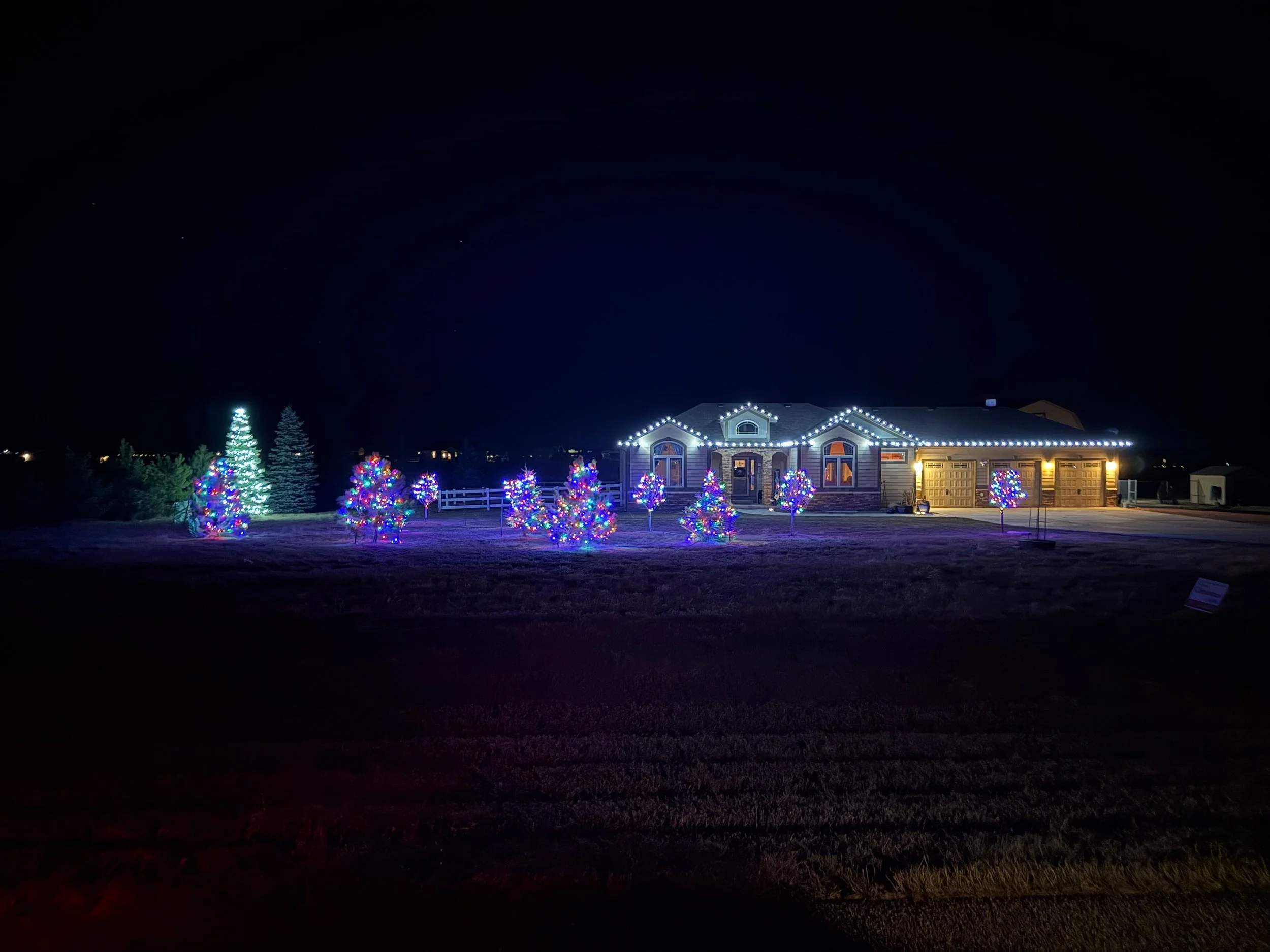 A house decorated with white Christmas lights outlining the roof, windows, and garage door, with colorful illuminated trees and a lit Christmas tree in the front yard at night.