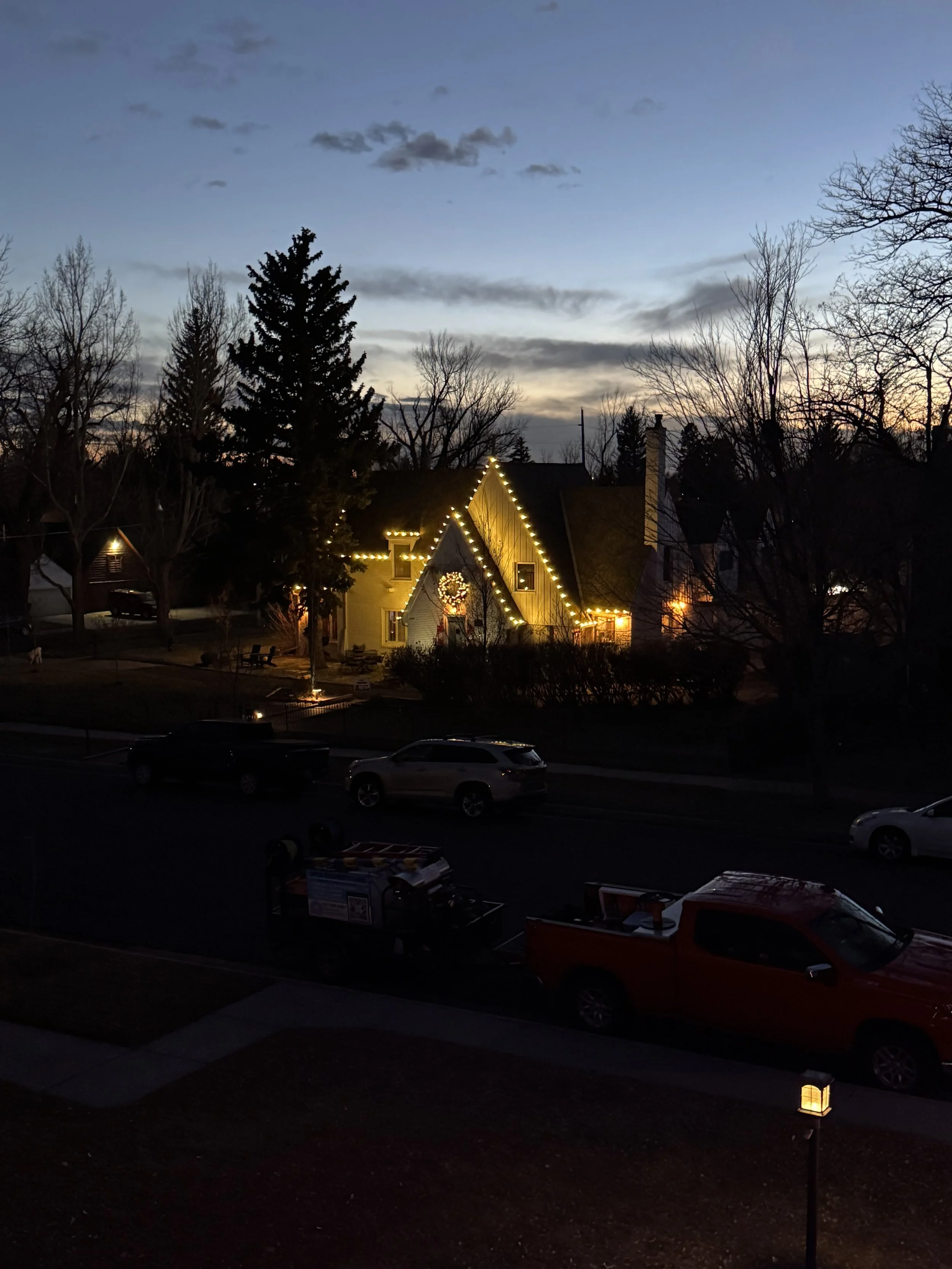 A house decorated with white Christmas lights and a wreath, surrounded by leafless trees, with a dark sky and some clouds at sunset.