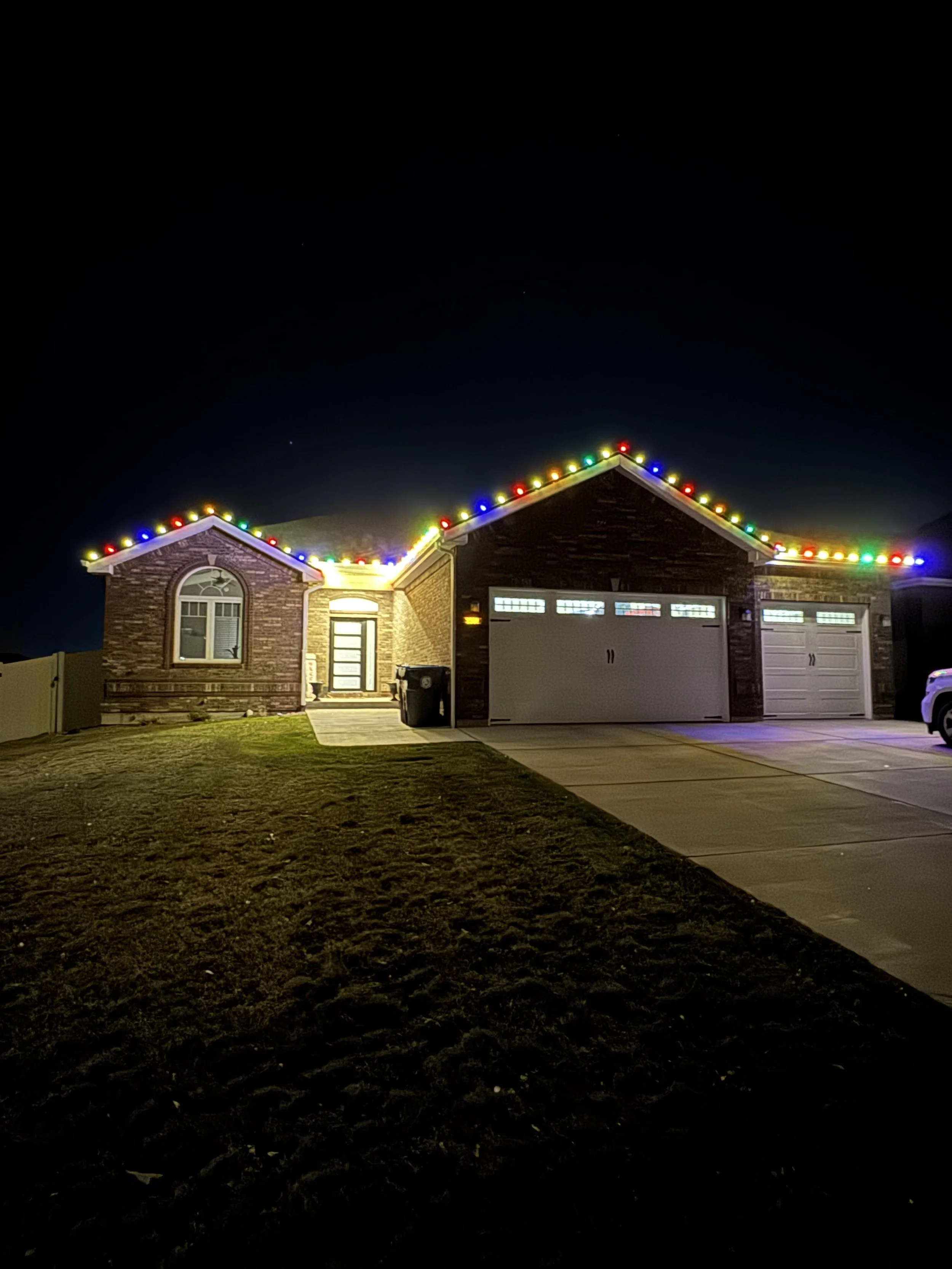 A house with string lights outlining its roof at night, two garage doors, a front door, a trash bin near the entrance, a lawn, and a dark night sky with a faint star.