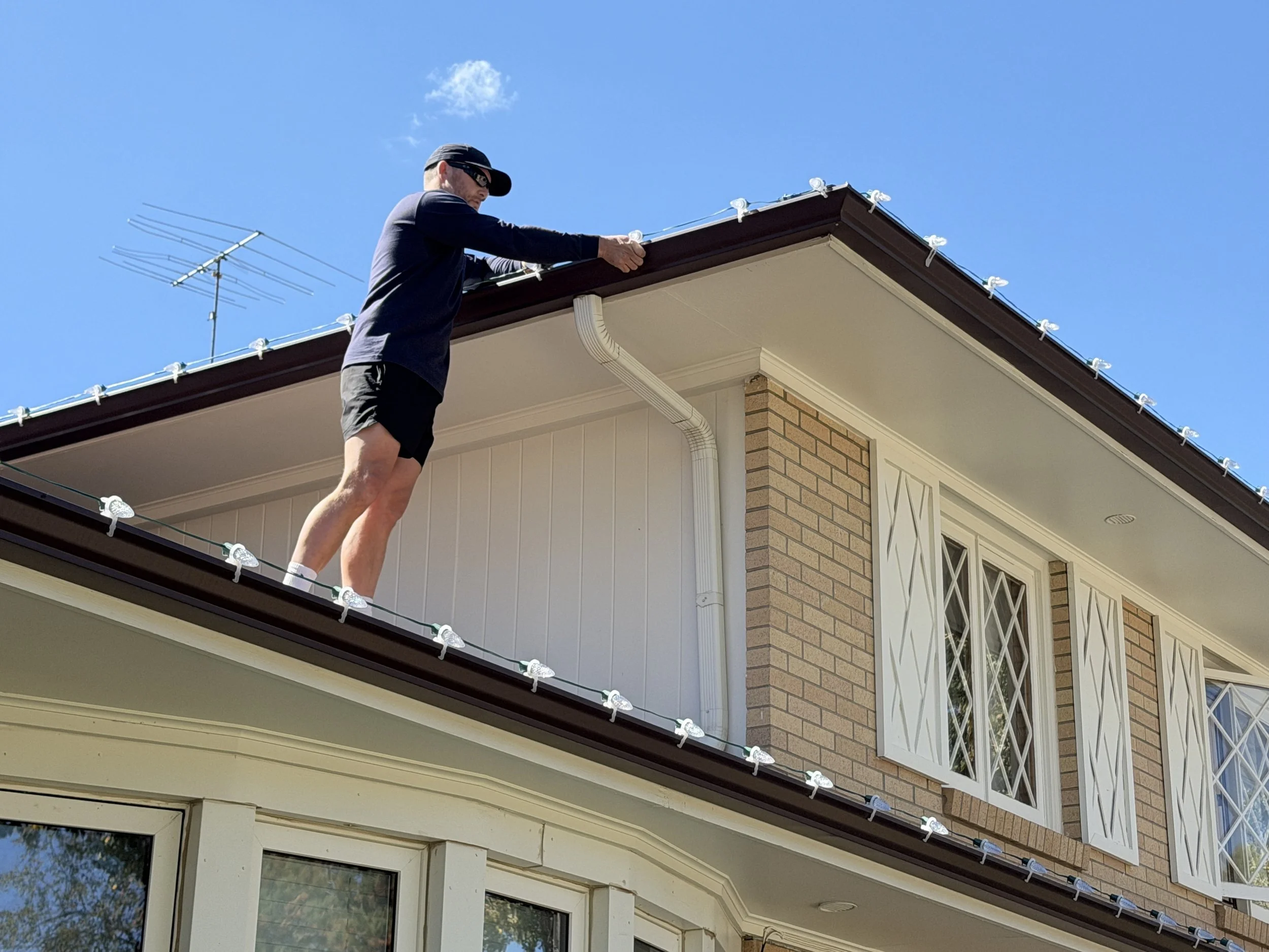 A man wearing sunglasses, a black cap, black shirt, and shorts is standing on the roof of a house, attaching Christmas lights under a clear blue sky.