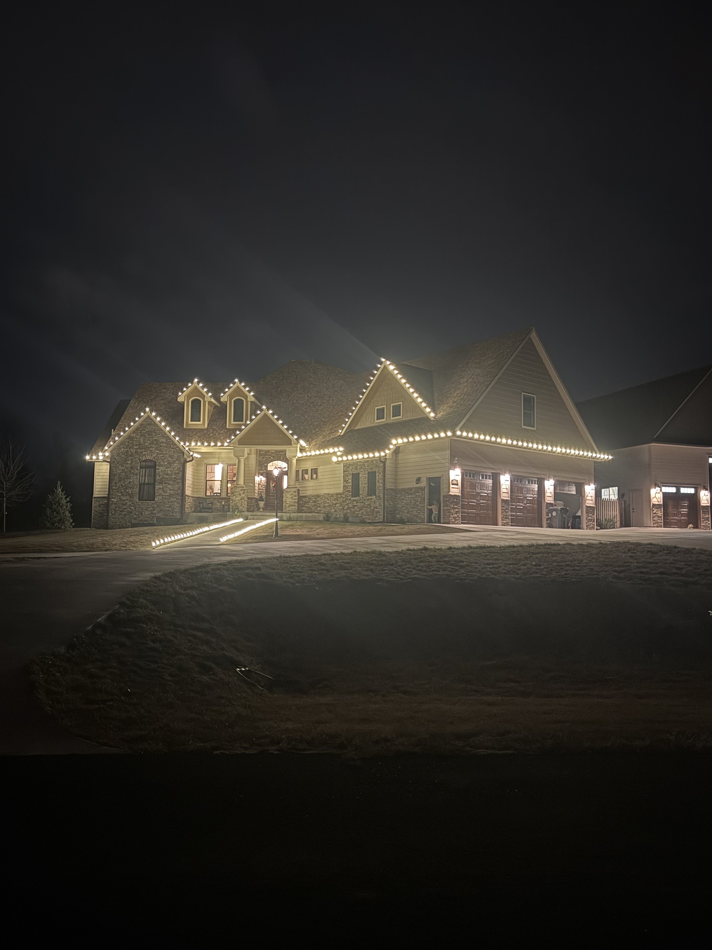 A house decorated with Christmas lights at night, with a large front yard and a driveway.