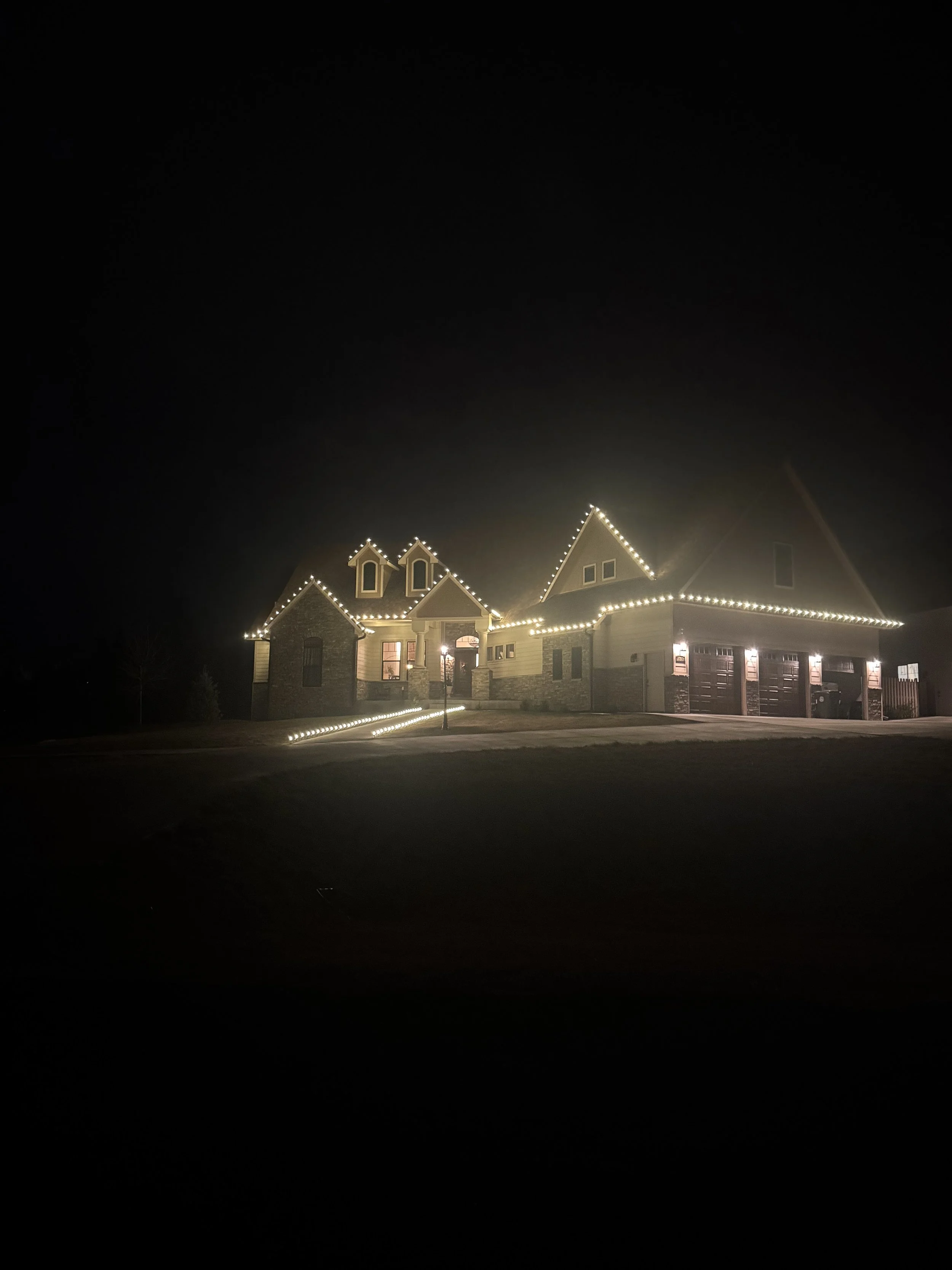 Nighttime exterior view of a house decorated with string lights outlining the roof, windows, and garage, with a well-lit driveway and a lamp post in the front yard.