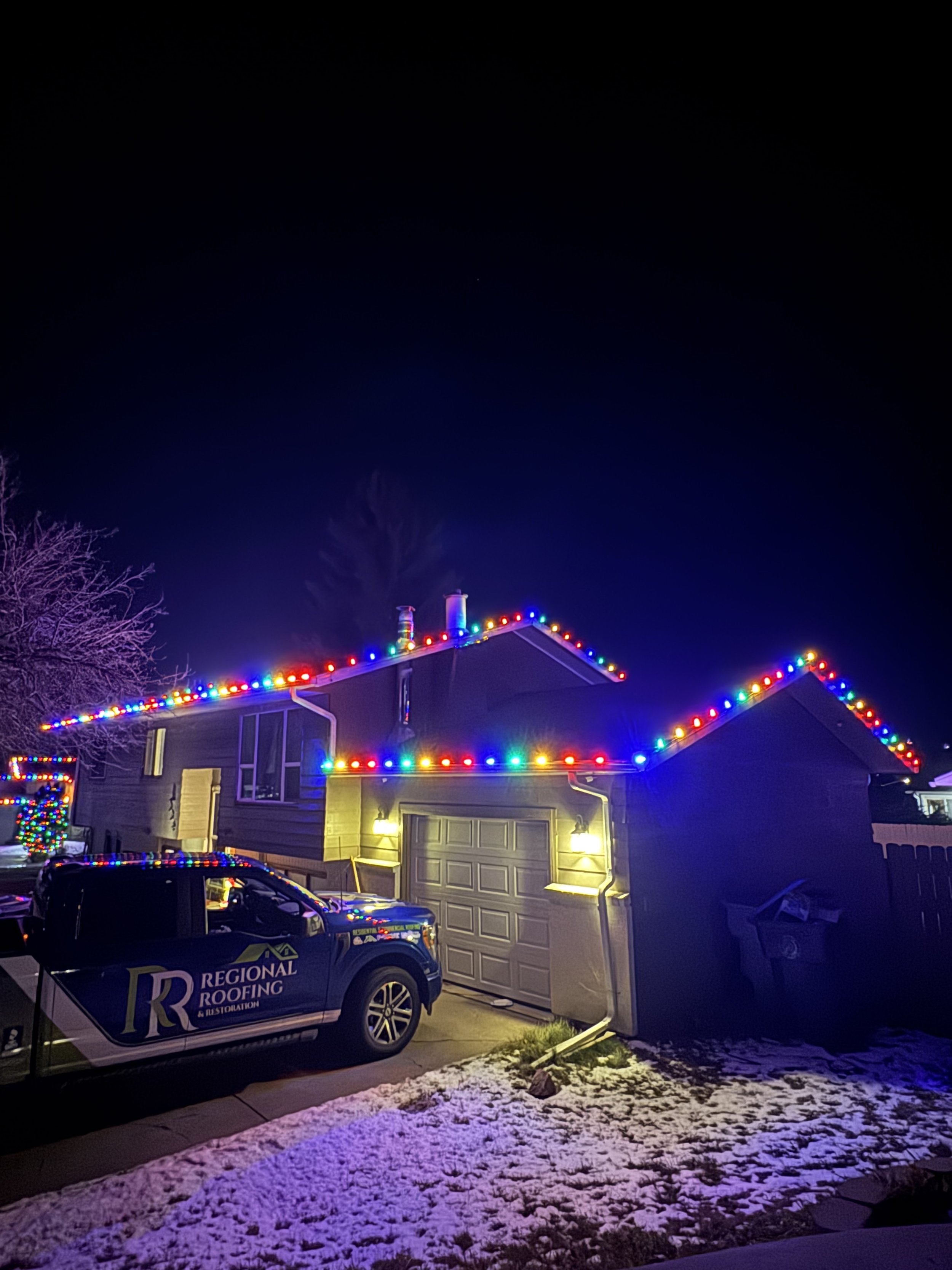 House decorated with colorful string lights at night, with snow on the ground and a vehicle parked in front.