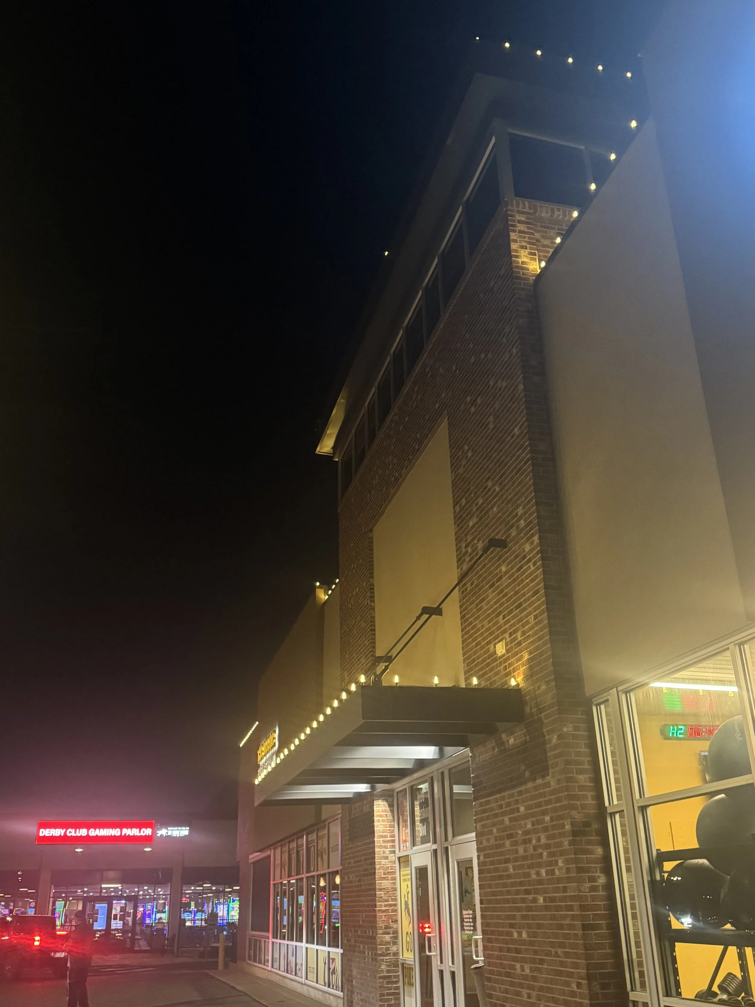 Nighttime view of a brick commercial building with lit signs, illuminated windows, and a parking lot, with a Casino sign in the background.