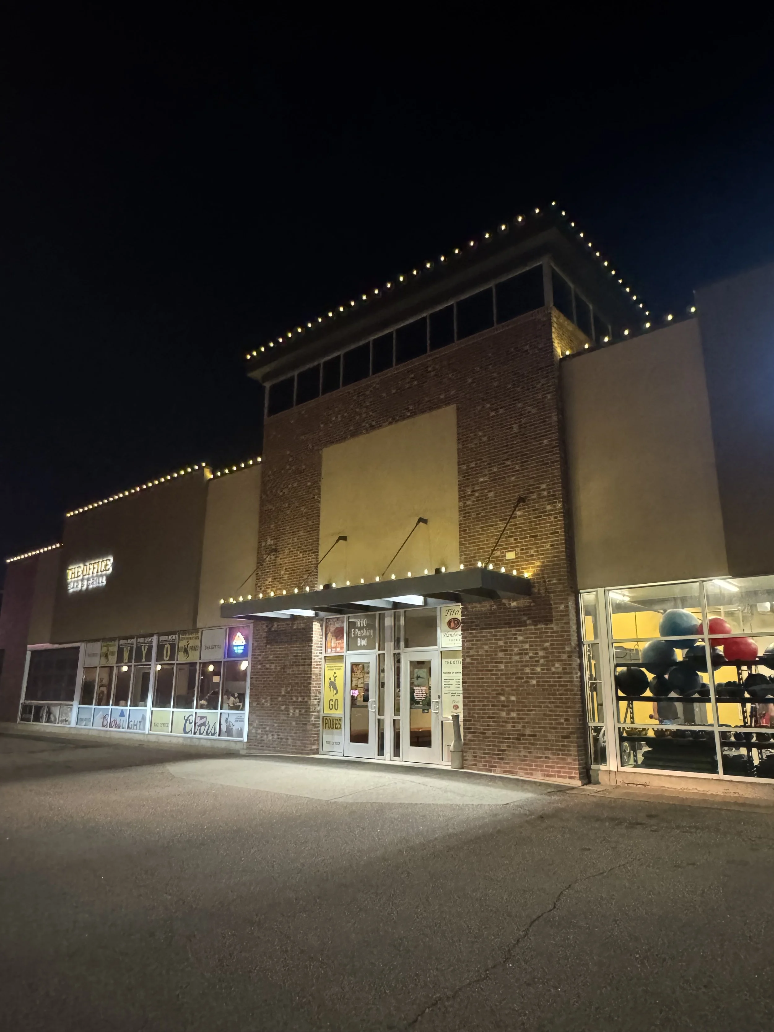 Night view of a brick building with large glass windows and an entrance door, decorated with string lights along the roofline, with a sign that reads 'THE OFFICE' and visible balloons on the right side.