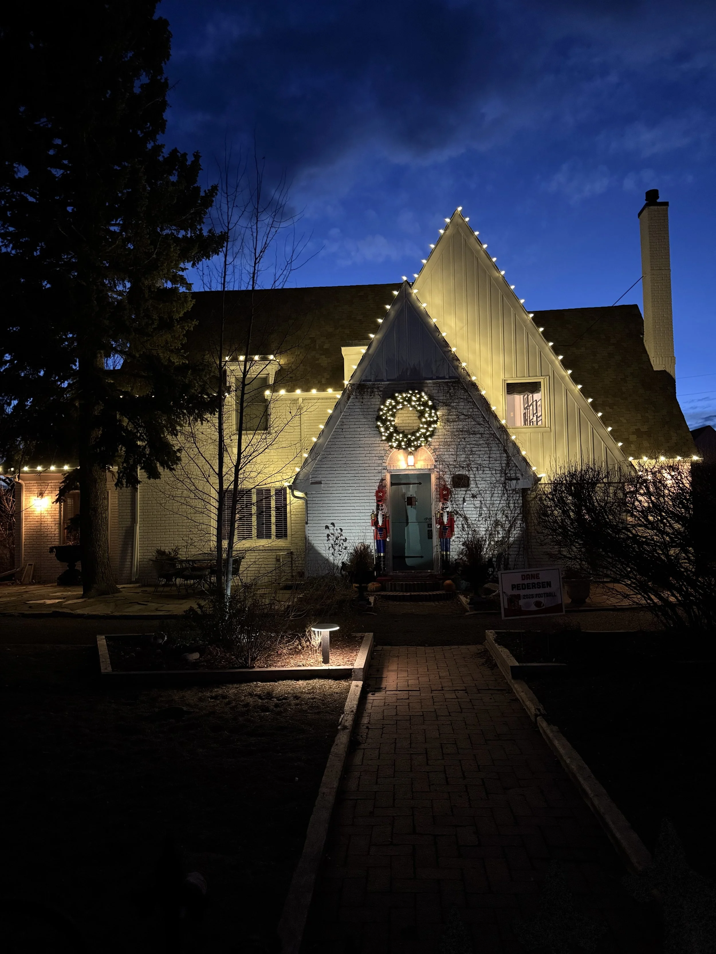 A house decorated with Christmas lights, a wreath, and Nutcracker figures, taken at dusk or night with a cloudy sky and trees in the surroundings.