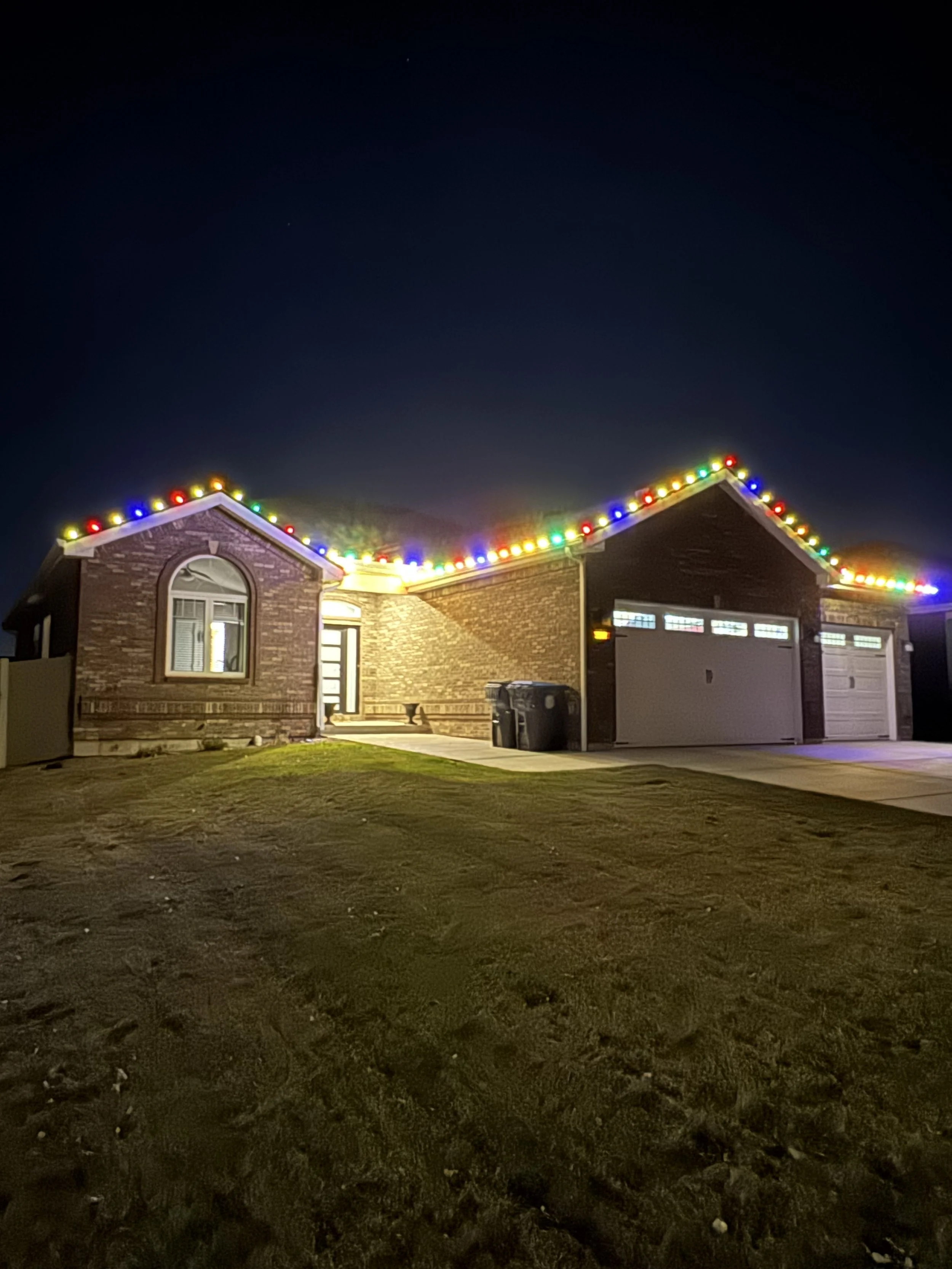 Nighttime photo of a house decorated with colorful holiday lights along the roofline, with a driveway and a garage, and some trash bins placed beside the house.