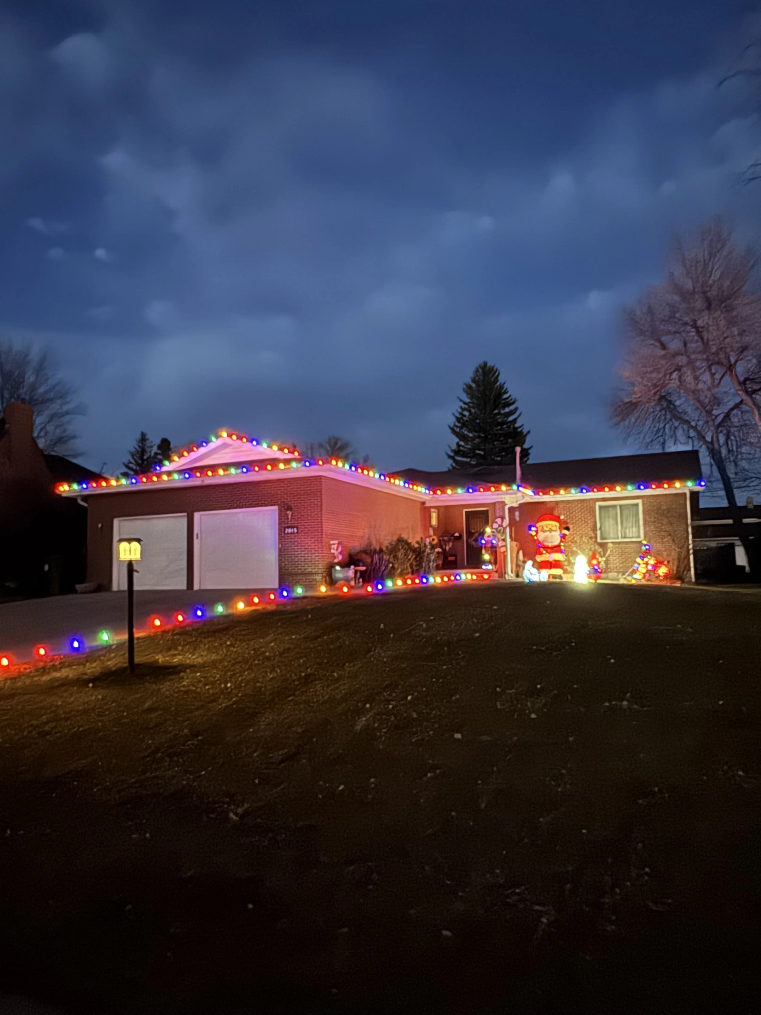A house decorated with colorful Christmas lights, with illuminated Santa Claus and Christmas trees outside, under a dark, cloudy sky.