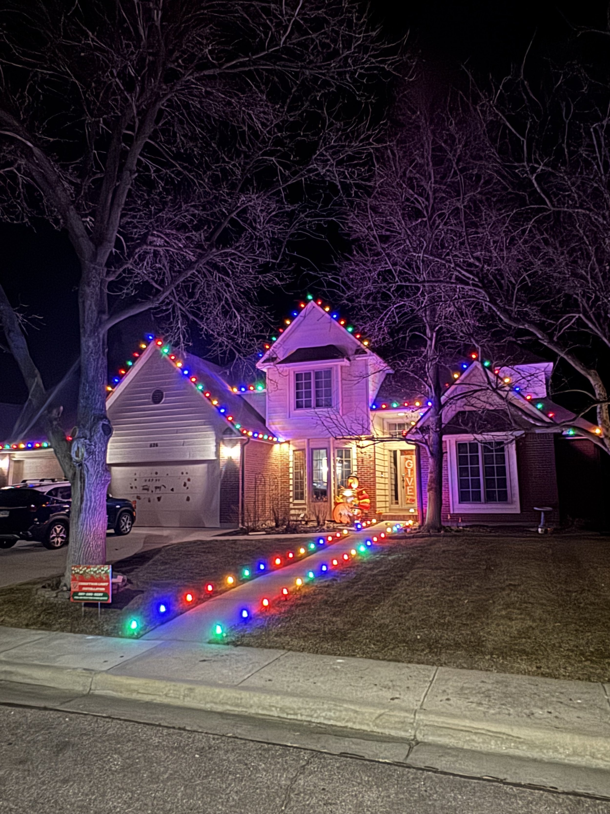 A house decorated with multicolored Christmas lights along the roof and walkway, with a large inflatable Santa Claus and Christmas ornaments in the front yard. Bare trees surround the house at night.