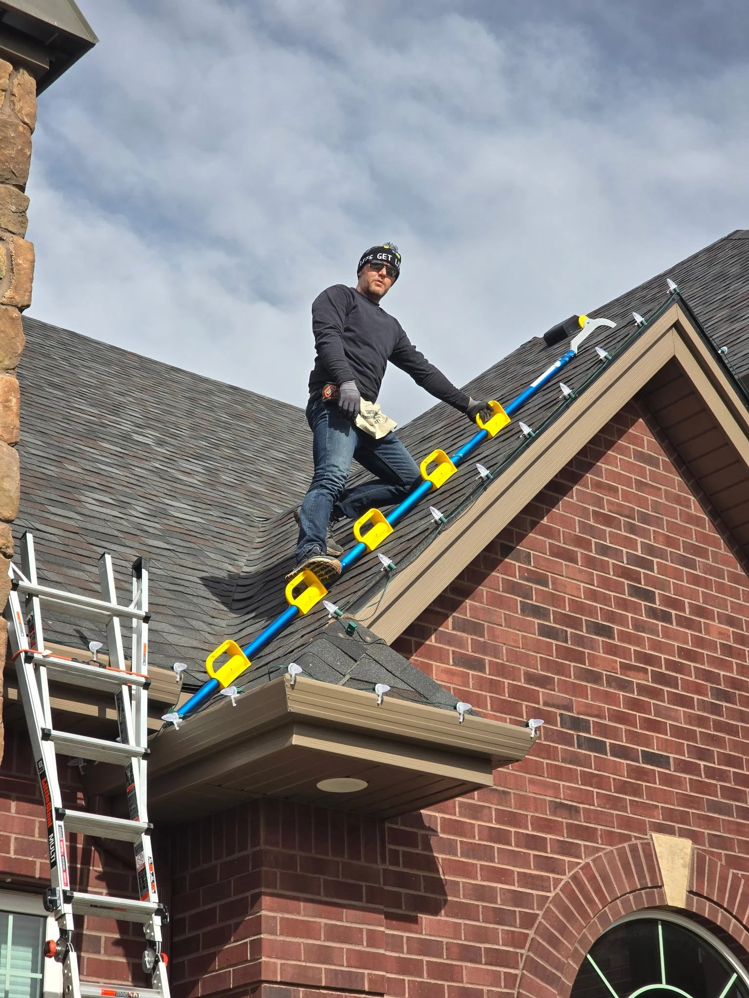 Man on a roof installing shingles, using a tool, with a ladder nearby, on a brick house with a large arched window, under a cloudy sky.
