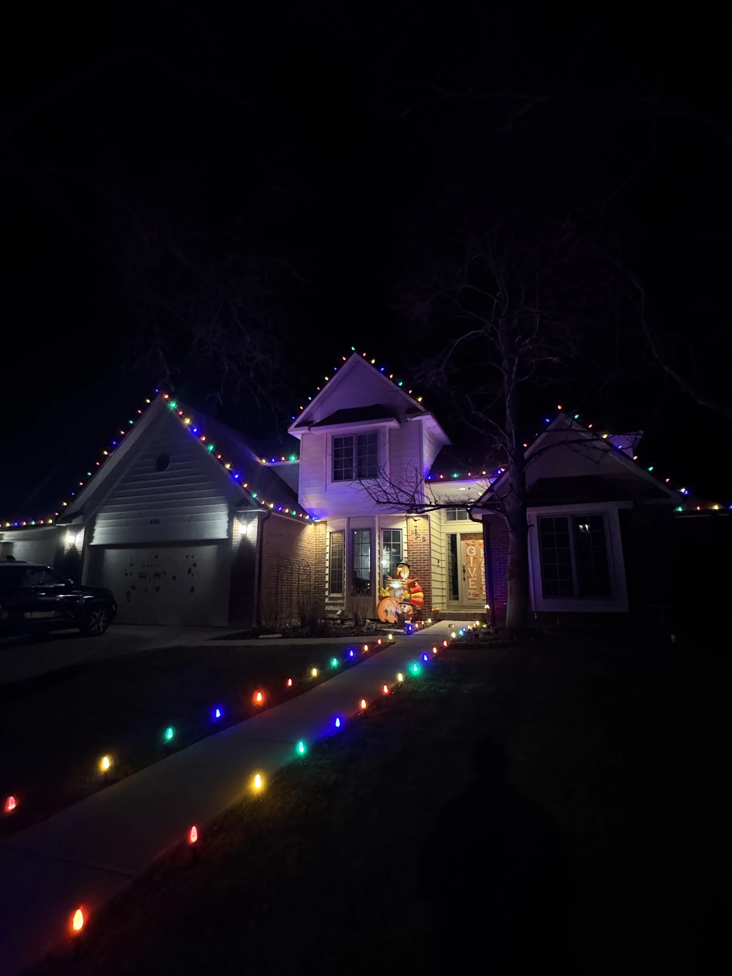 A house decorated with colorful Christmas lights along the roofline and the garden pathway, with a festive inflatable decoration in front of the house at night.