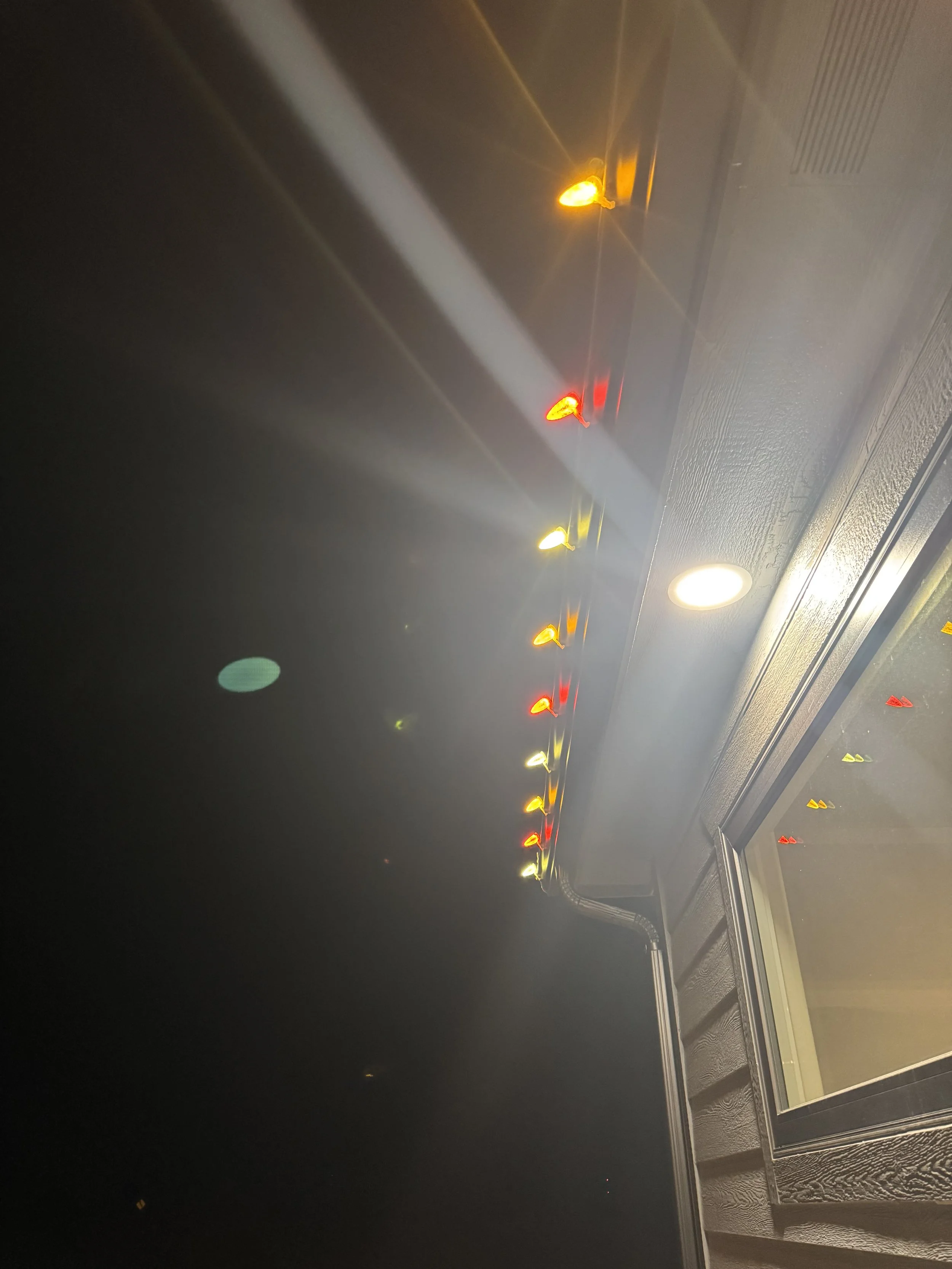 Ceiling with a row of yellow, red, and white decorative LED lights reflected in a mirror, along with a ceiling vent, on a painted dark wall.