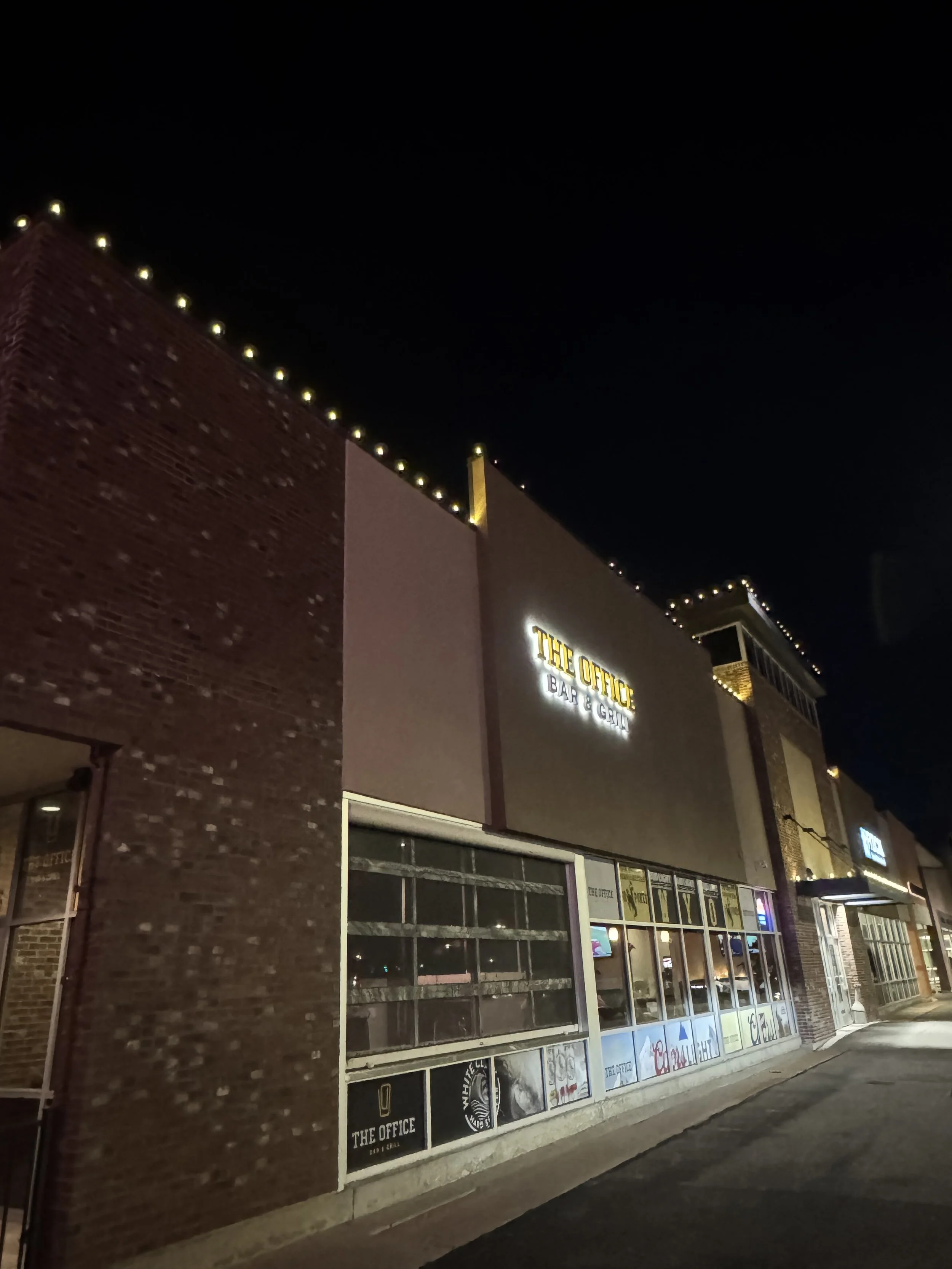 Night view of a building with illuminated sign reading 'The Office Bar & Grill' and decorated with string lights along the rooftop.