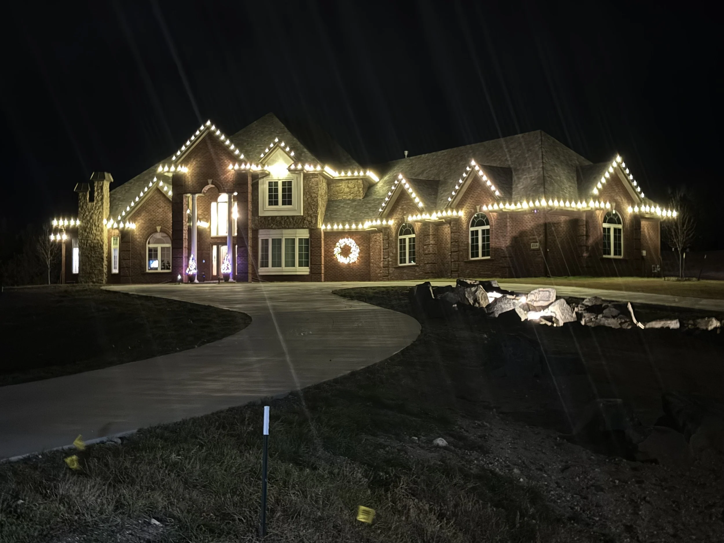 A house decorated with Christmas lights at night, with a wreath on the front wall and illuminated Christmas trees near the entrance, and a landscaped yard with rocks and a curved driveway.