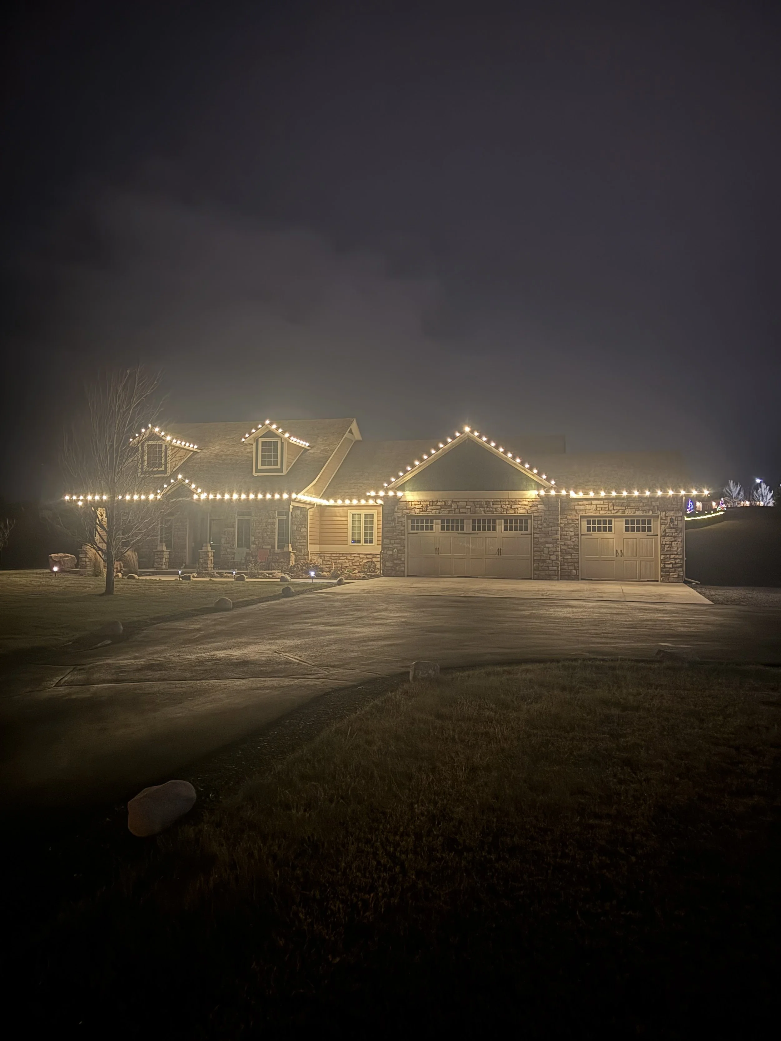 A house decorated with Christmas lights at night. The house has a stone facade and a garage with three doors. String lights outline the roof edges. The front yard has a tree and some rocks, with a driveway leading to the garage.