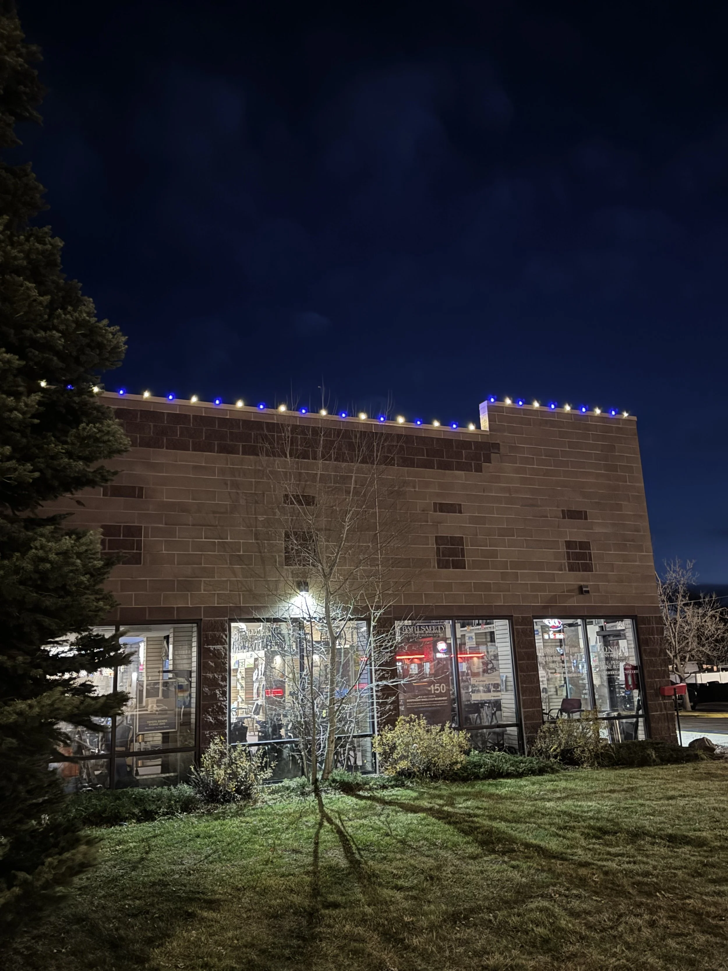 Nighttime view of a brick building with large glass windows, illuminated interior, and a small grassy area with trees and bushes in front. The building has decorative blue and white string lights along the roofline.