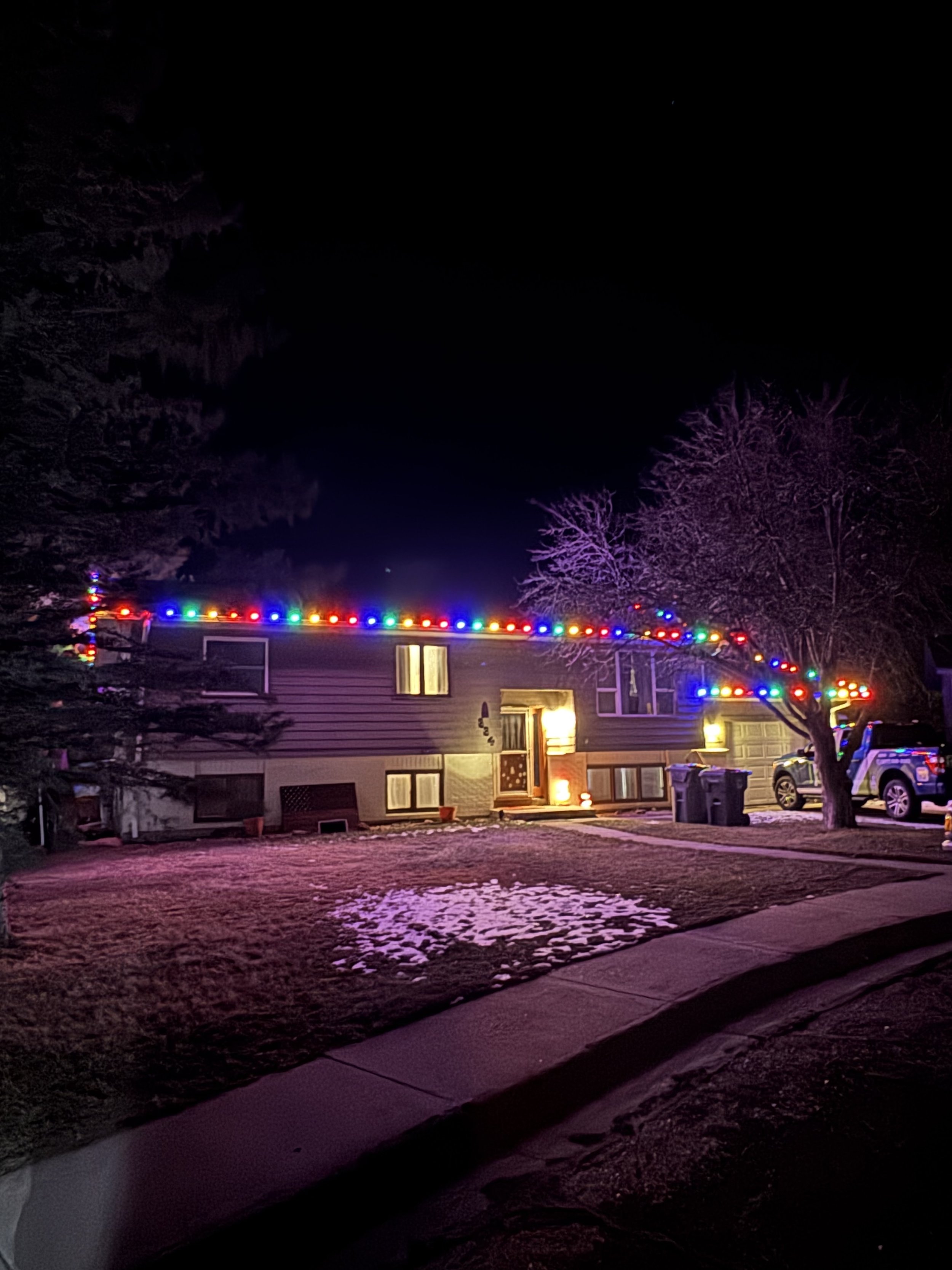 A house decorated with colorful Christmas lights, with a snow-covered front yard and trees, at night.