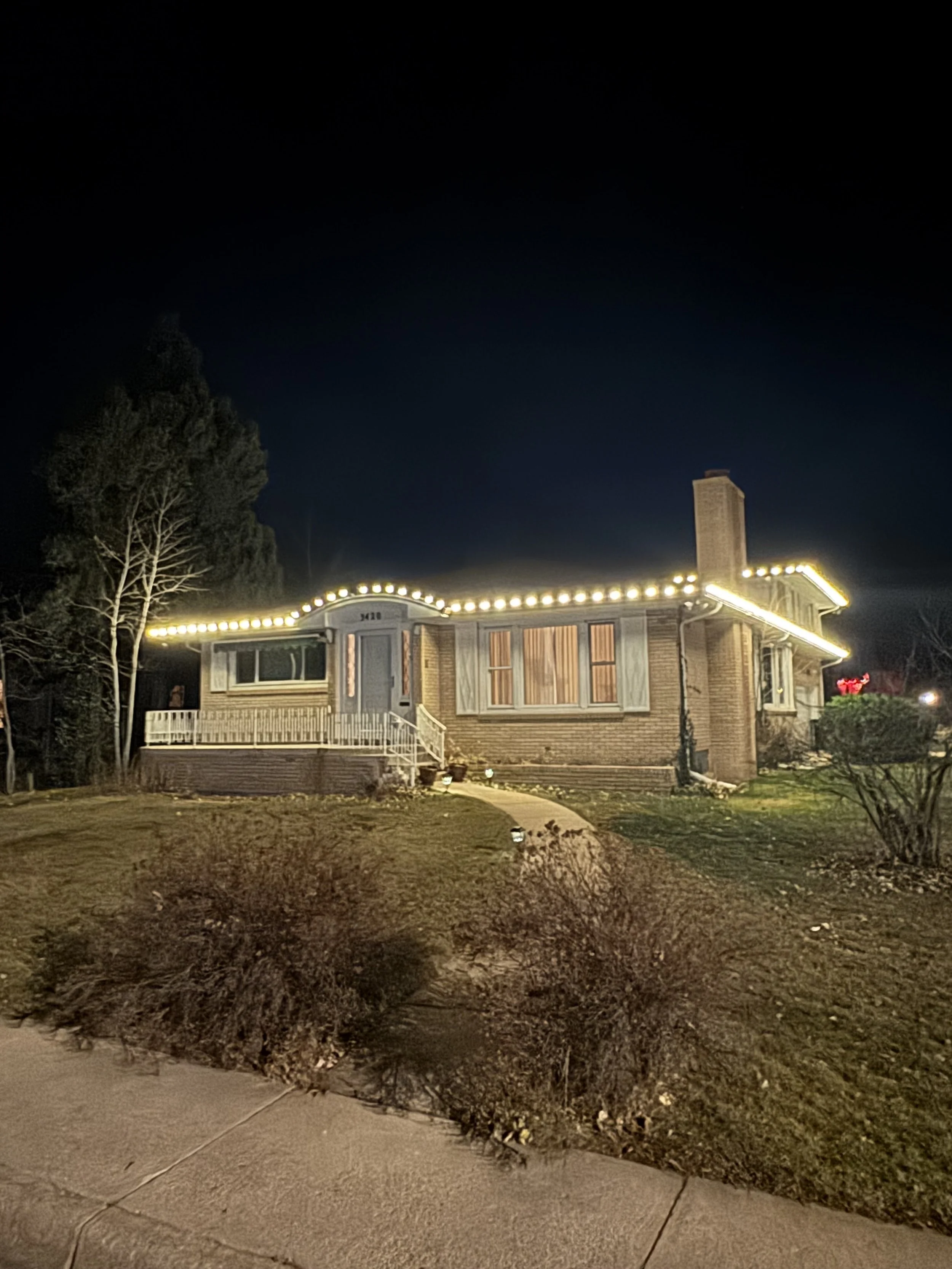 A house decorated with string lights at night, with trees and a pathway in the yard.