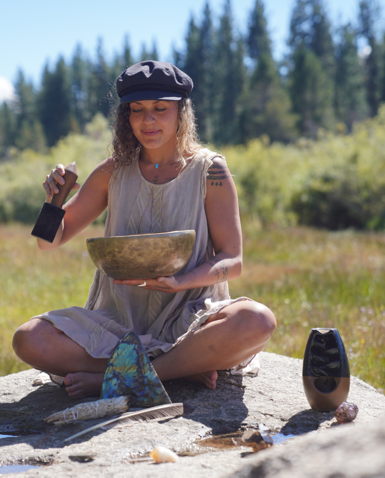 A woman with curly blonde hair, wearing a black cap and a sleeveless beige dress, sits cross-legged outdoors in a natural setting with trees in the background. She is holding a singing bowl and a mallet, with a relaxed and peaceful expression. In front of her are two other singing bowls, one eye-shaped and one round, along with decorative feathers and crystals, suggesting she is practicing sound healing or meditation.