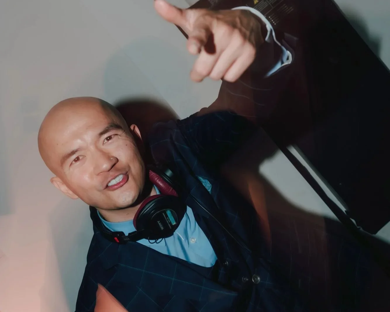 A man with a bald head and headphones around his neck pointing upward with his finger, smiling at the camera.