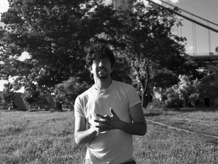 A man with curly hair, glasses, and a beard standing outdoors in a grassy park with trees and a bridge in the background, wearing a light-colored t-shirt.