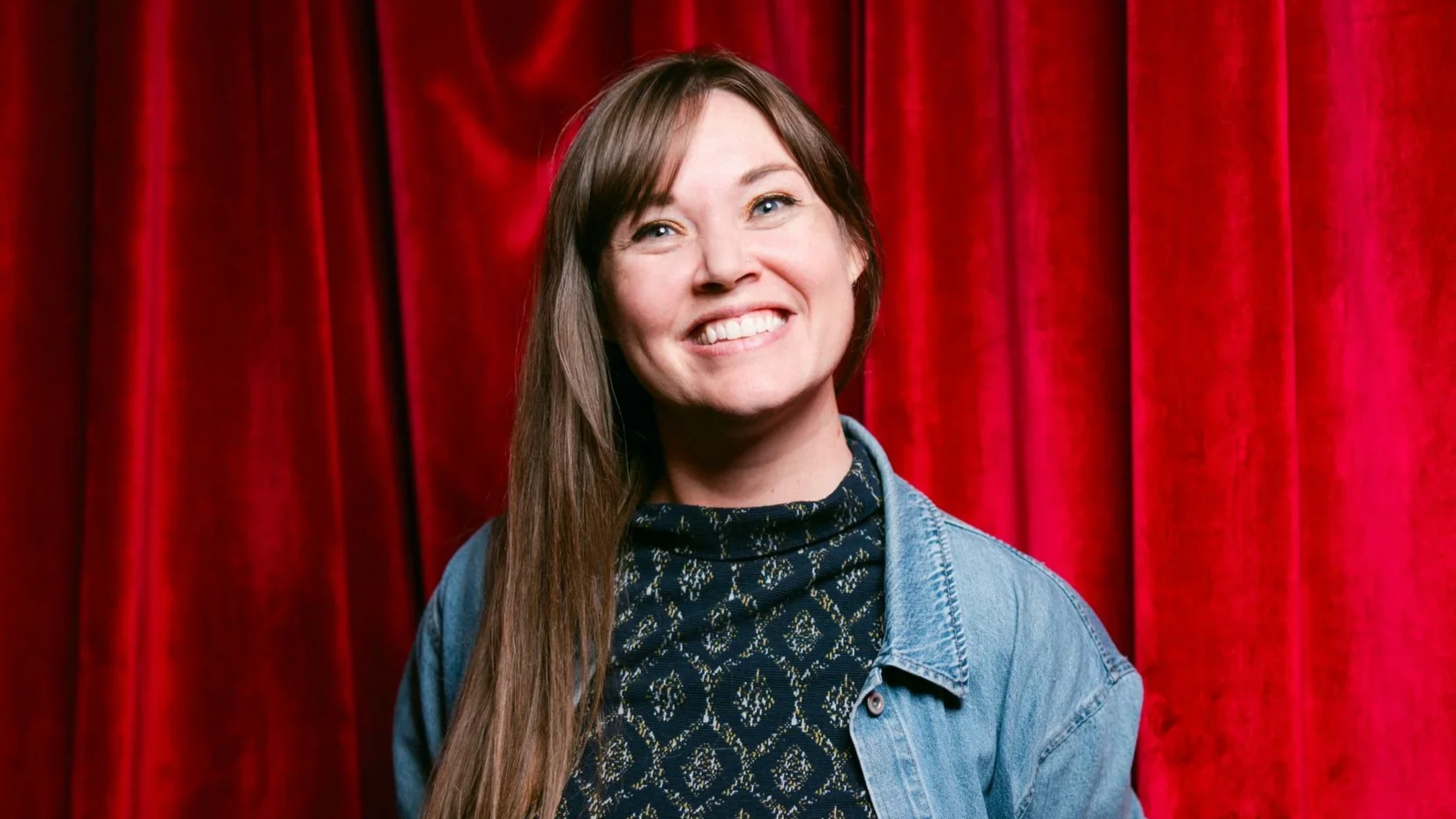 A smiling woman with long brown hair standing in front of red velvet curtains.