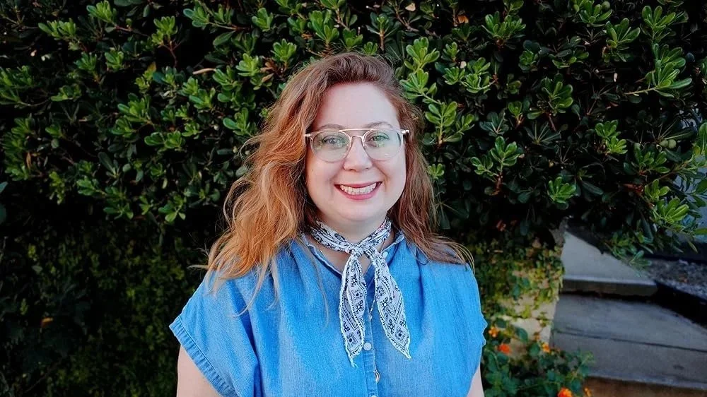 Smiling woman with glasses and wavy, shoulder-length red hair wearing a blue blouse and patterned scarf, standing outdoors in front of lush green foliage.